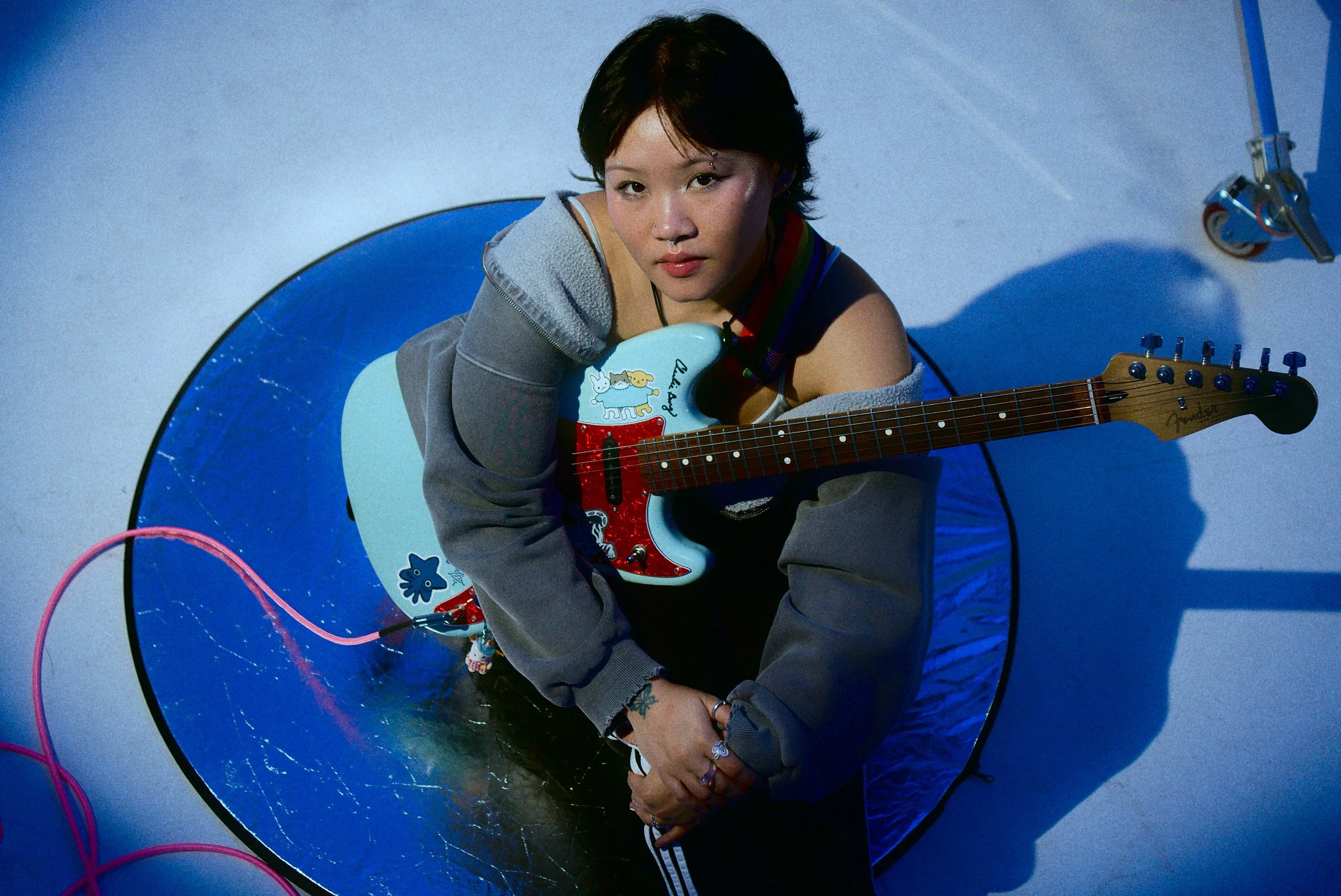 Young woman sitting on the floor holding a light blue electric guitar with cartoon stickers, wearing a gray hoodie off the shoulder, in a studio with a blue backdrop and a reflector, casting a shadow.
