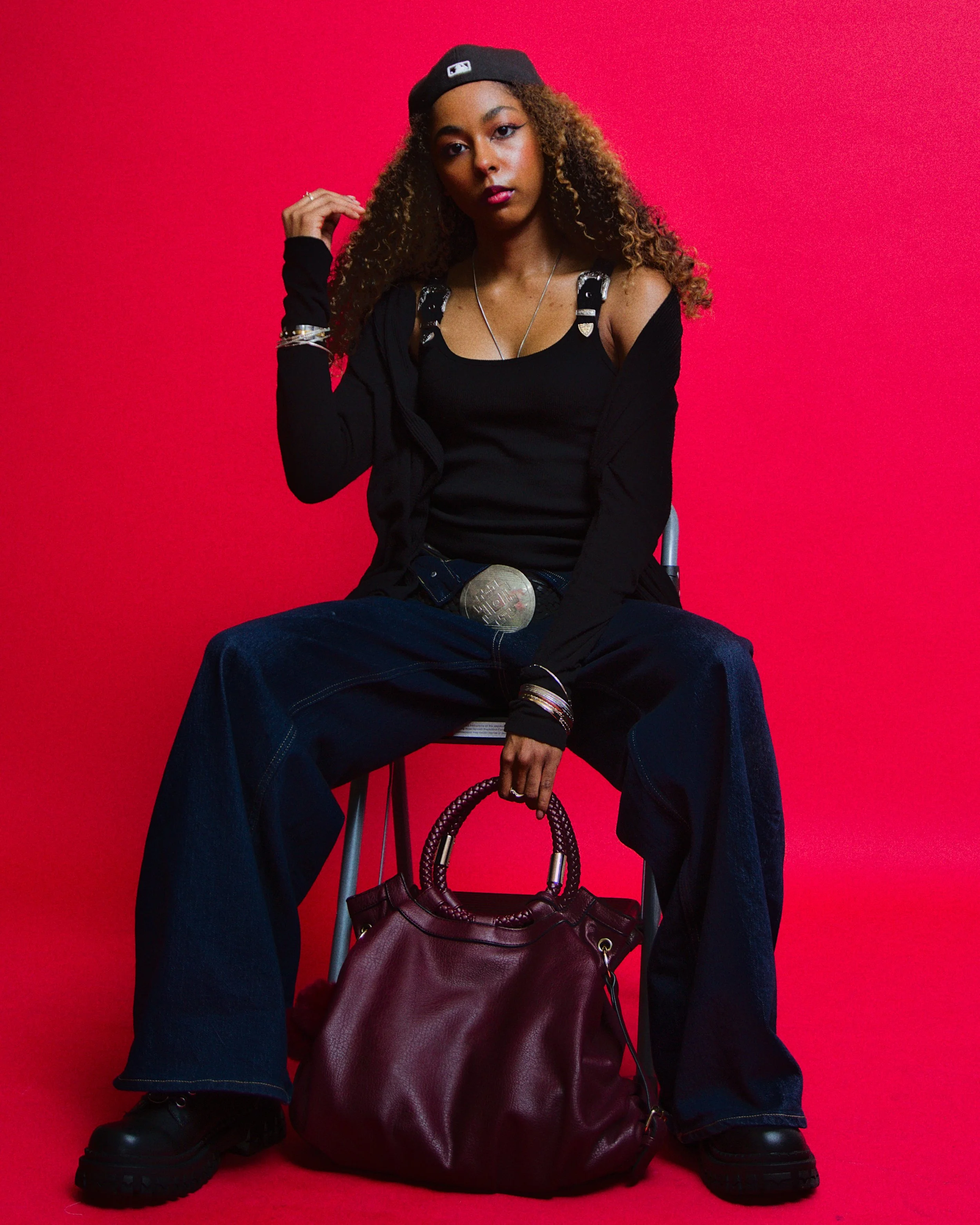 A young woman with curly hair sitting on a chair against a red background. She is wearing a black tank top, a black jacket with shoulder embellishments, baggy jeans, black boots, a backward black cap, and accessorized with bracelets, necklaces, and a