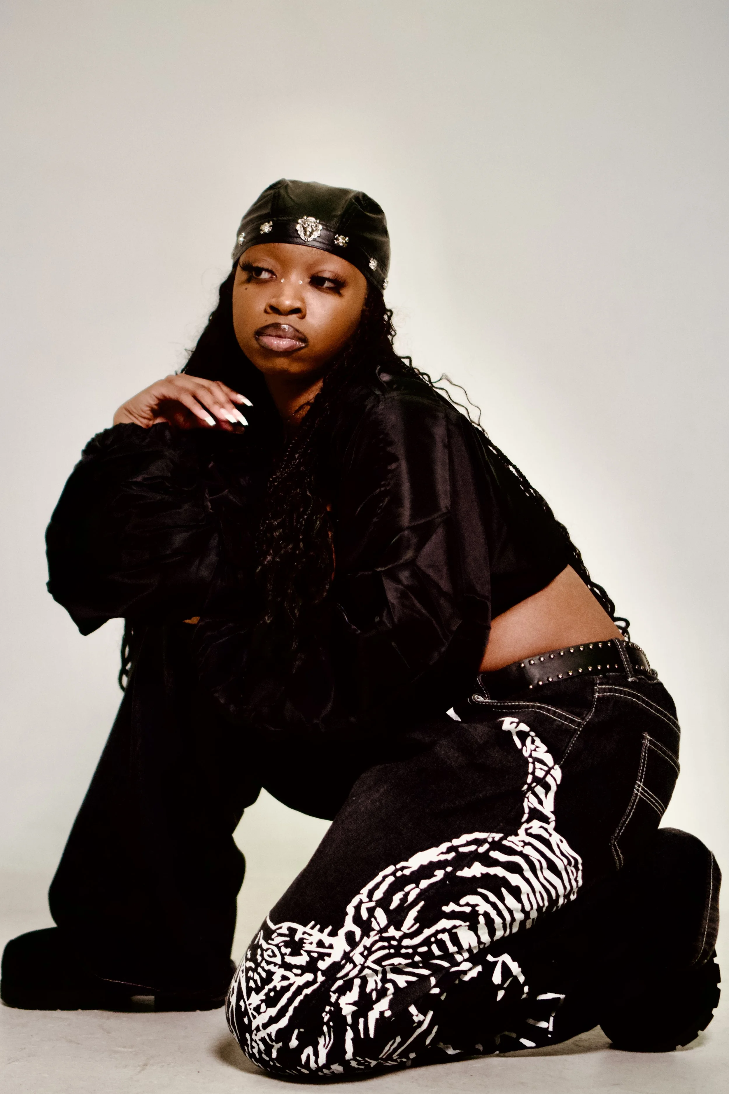 A woman with long braids wearing a black satin jacket, black pants with white patterns, a black leather cap with embellishments, and a studded belt, kneeling on the floor with one hand near her face against a plain background.