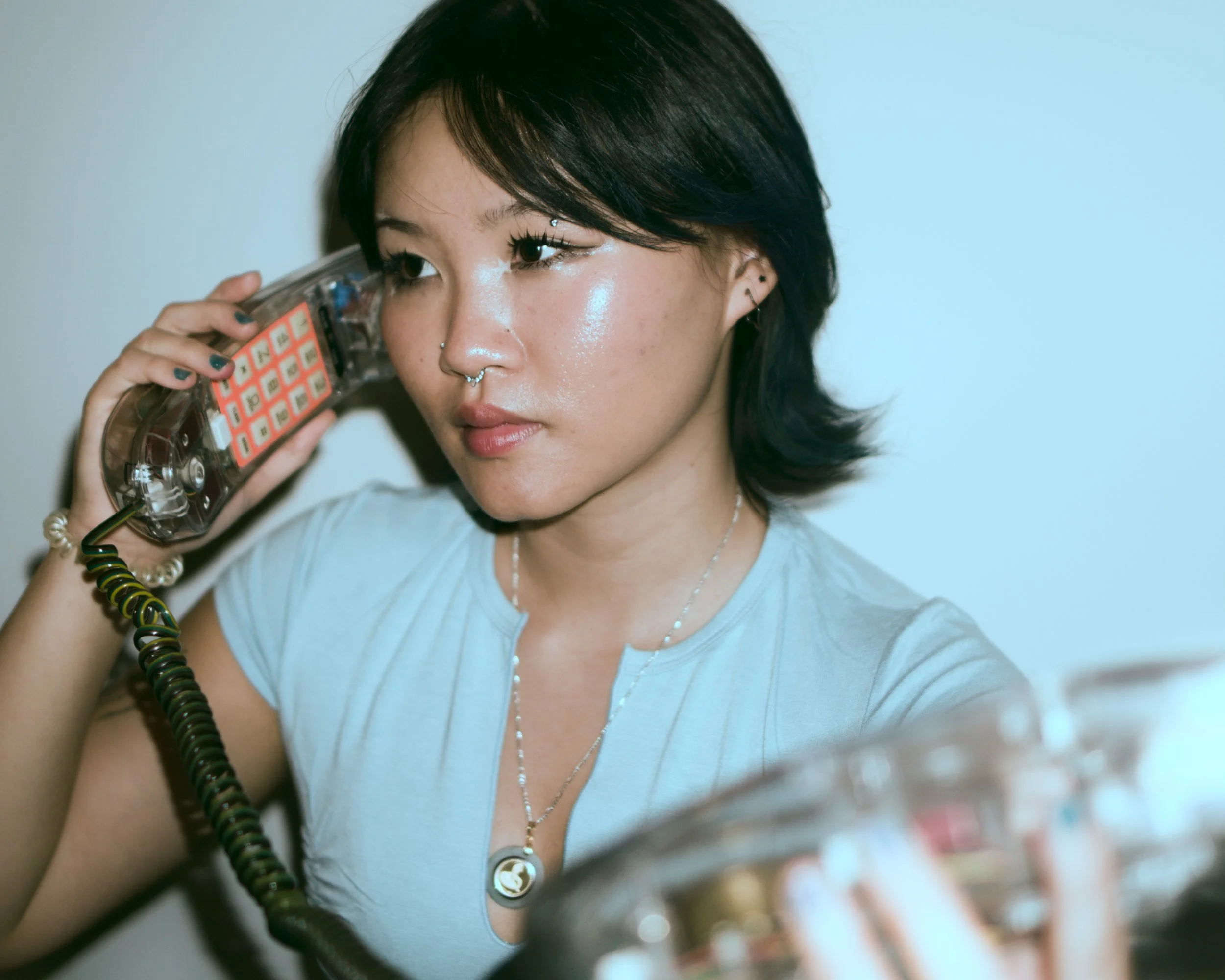 Nina1am with short dark hair, tan skin, and multiple piercings, holding a retro phone to her ear, while looking at something off-camera. They are  wearing a light blue shirt, a necklace, and a bracelet.