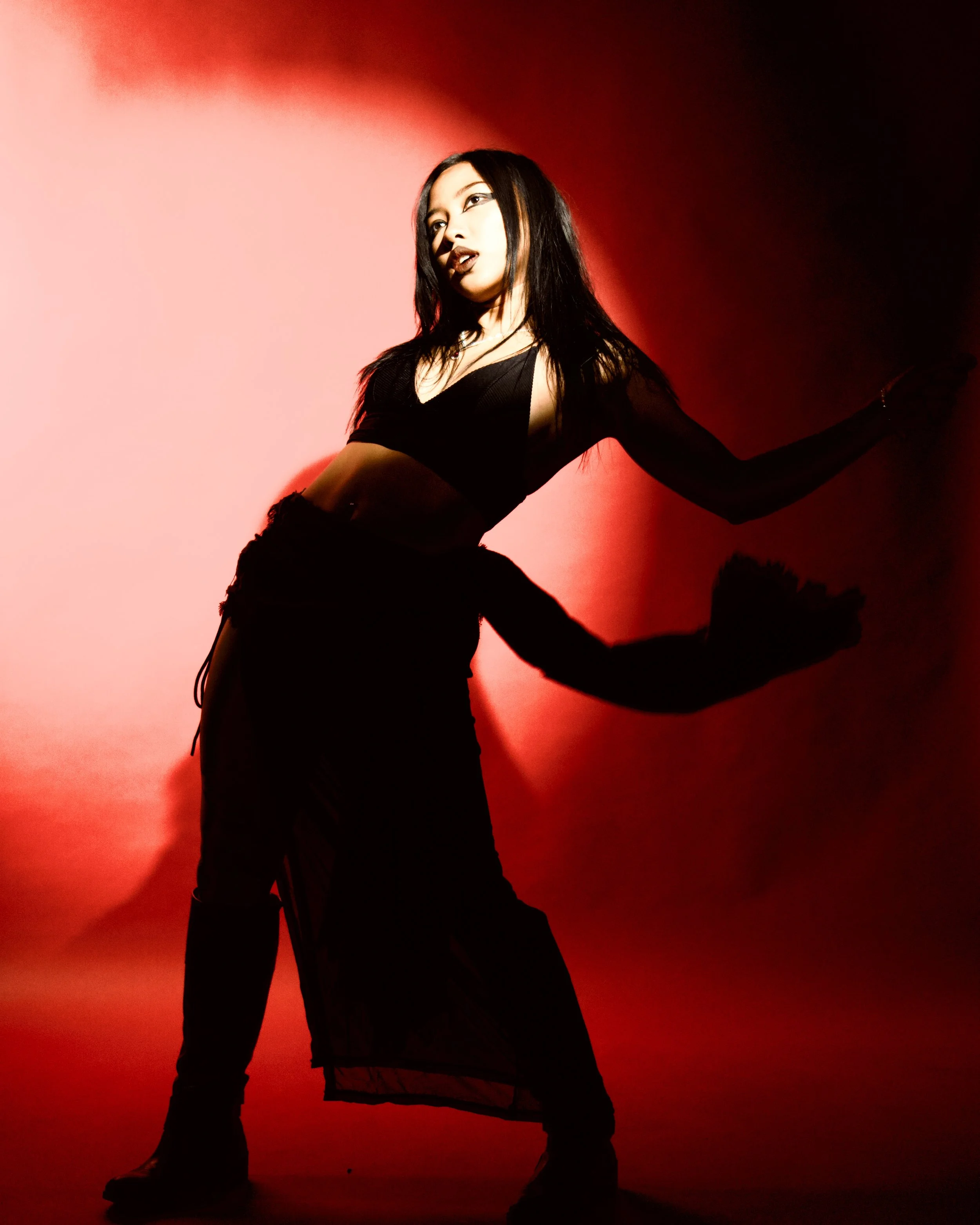 A woman with long black hair posing against a red gradient background, dressed in black. She is standing with one arm extended and looking to the side. Photographed by Erin De Shay