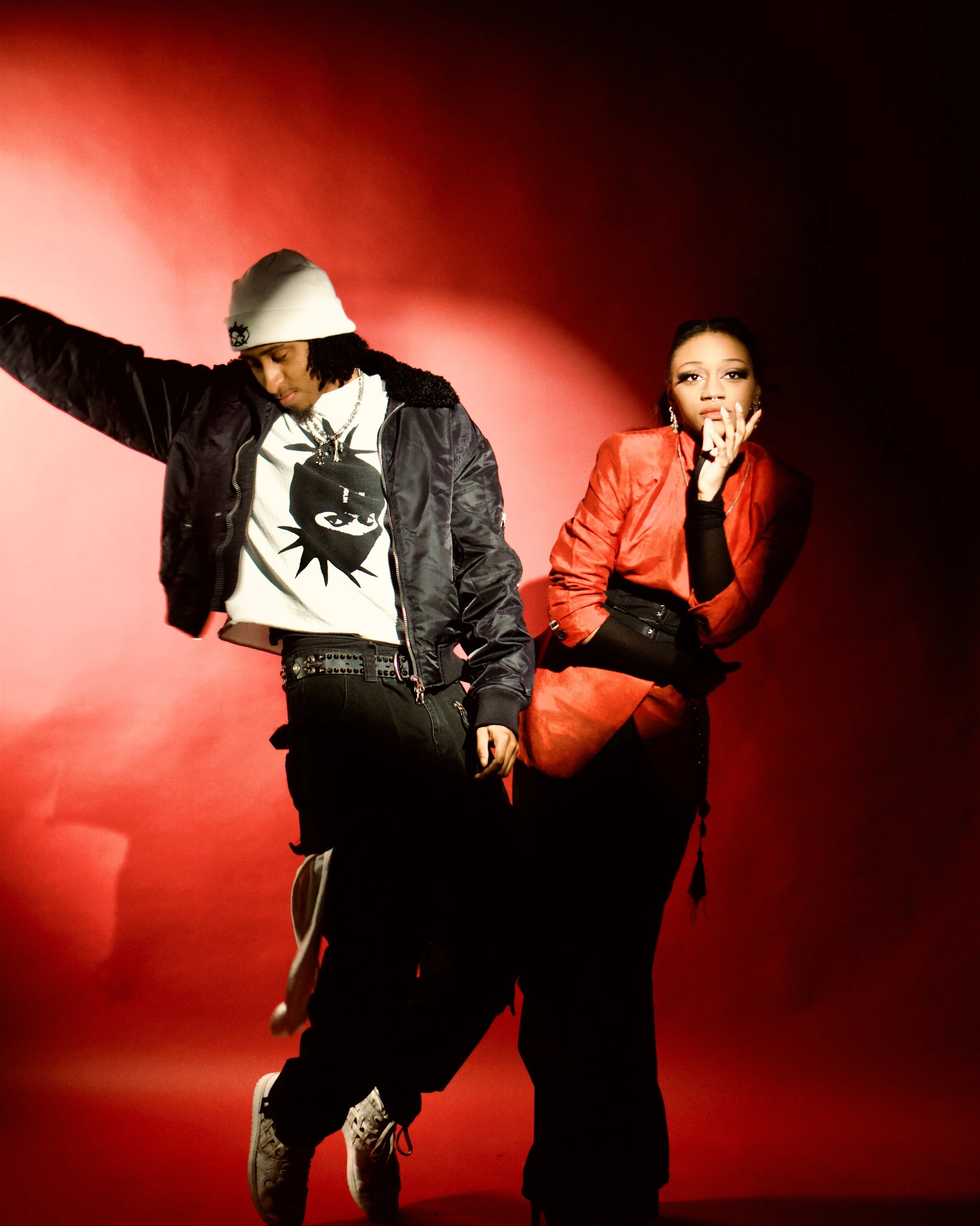 Two young adults pose against a red and black background, with dramatic lighting highlighting their stylish streetwear outfits.  Photographed by Erin De Shay.