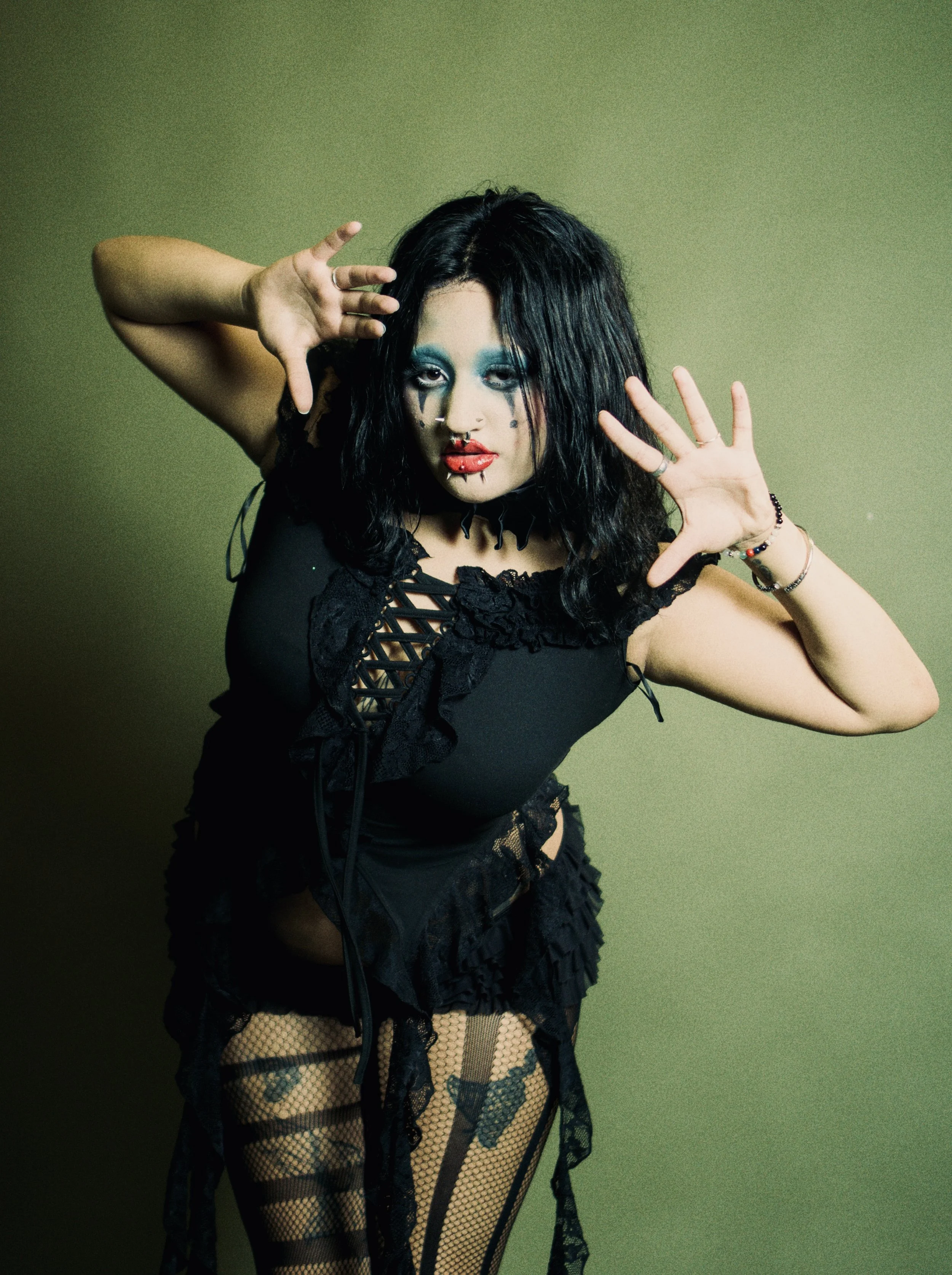 A woman with black hair and bold makeup, wearing black lace and fishnet clothing, striking a dramatic pose against a plain green background.  Photographed by Erin De Shay.