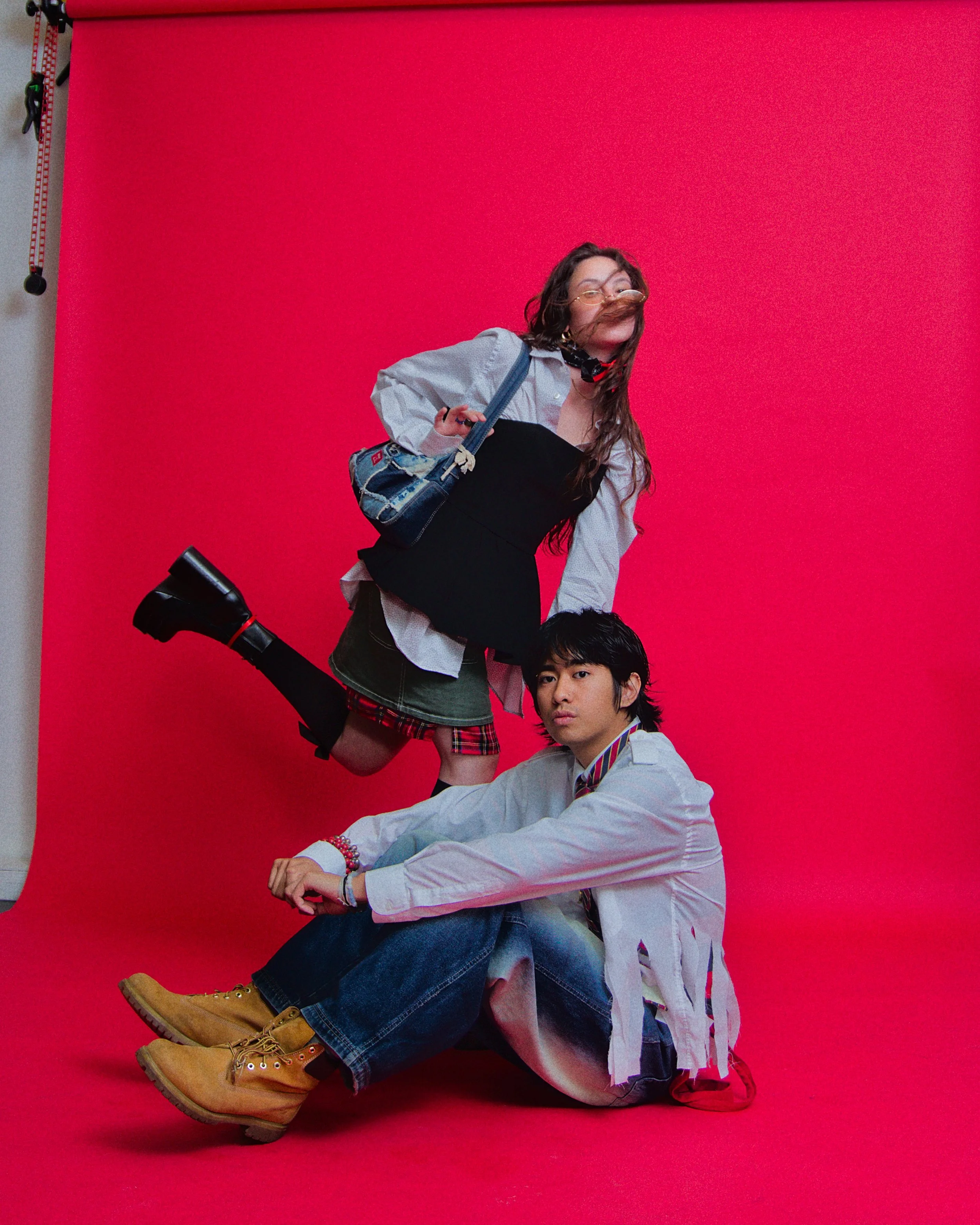 A young woman with glasses and long hair, wearing a black dress over a white shirt, black boots, and carrying a handbag, is posed mid-air against a red backdrop. A young man with dark hair, wearing a white jacket, jeans, and tan boots, is sitting on 
