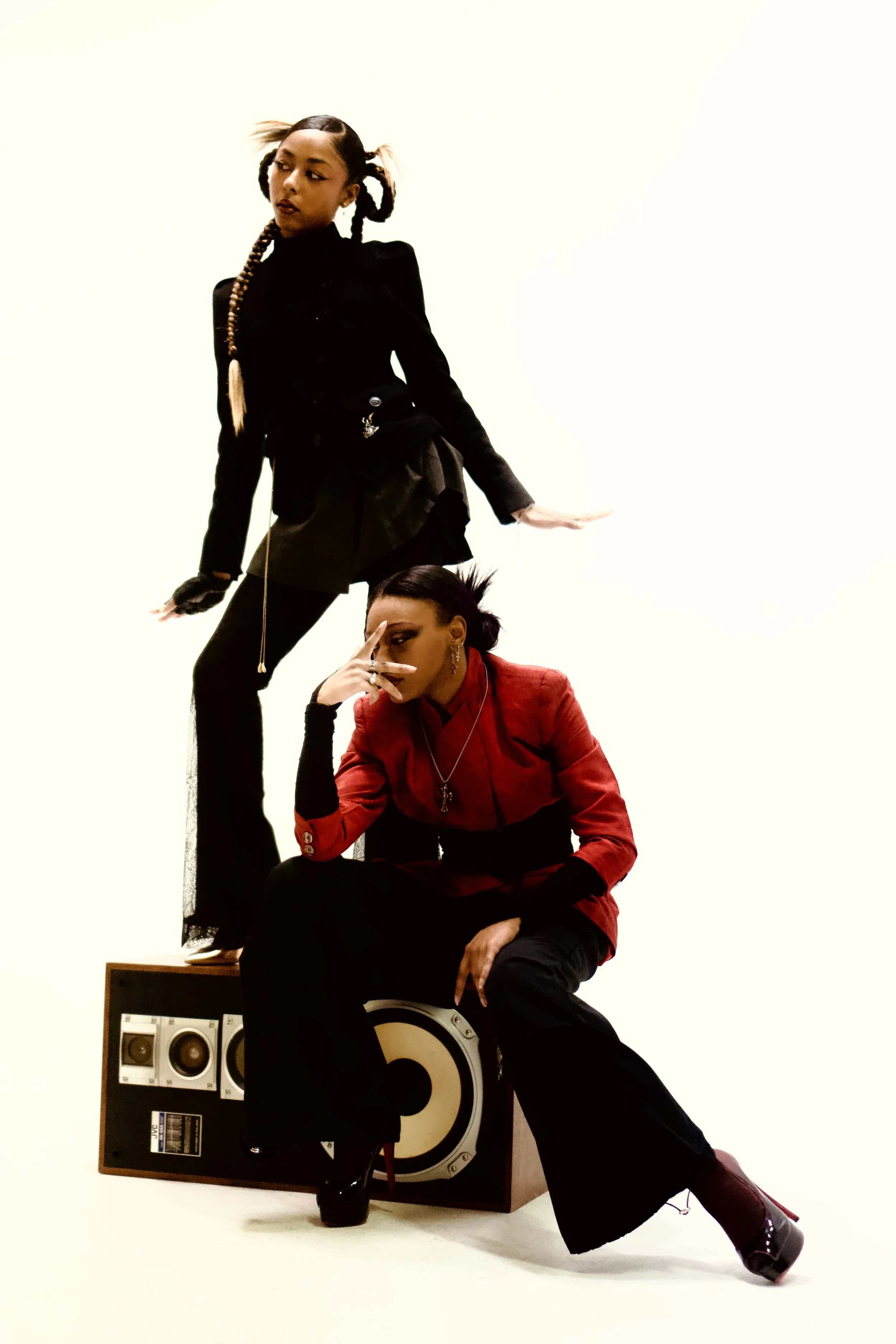  Two stylish women posing with a vintage speaker against a minimal background; one is standing on the speaker with a contemplative expression, and the other is sitting on it making a finger gesture near her face.  Photographed by Erin De Shay.