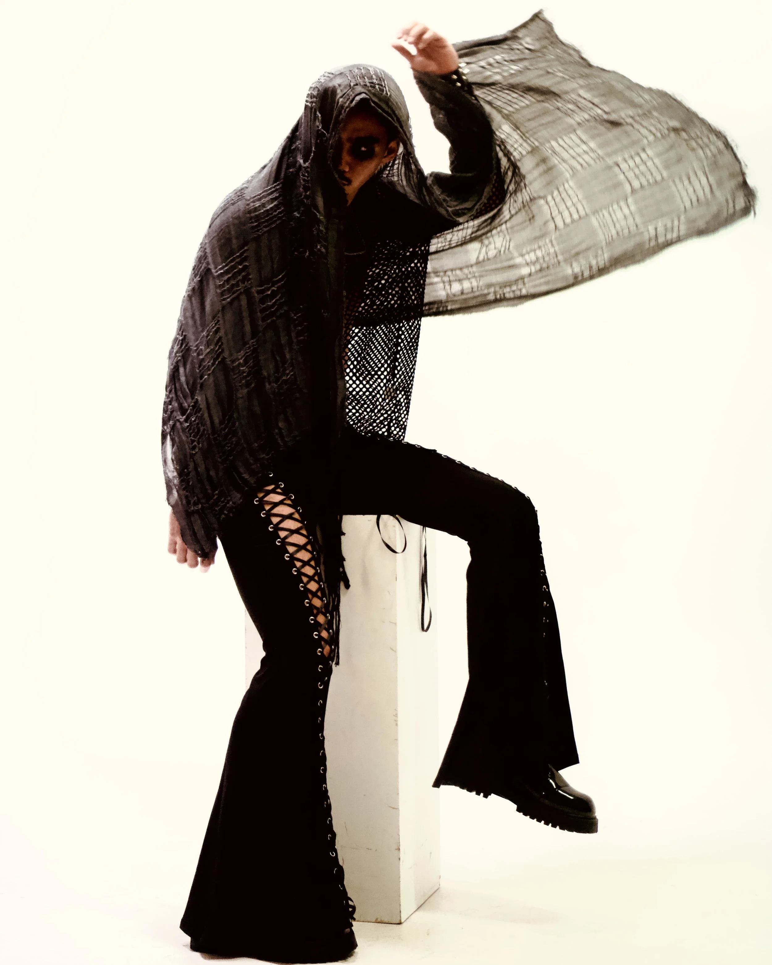 Person wearing a black lace-up pant with large platform shoes, sitting on a white cube, with a black lace shawl draped over the head and body, against a plain white background. Photographed by Erin De Shay