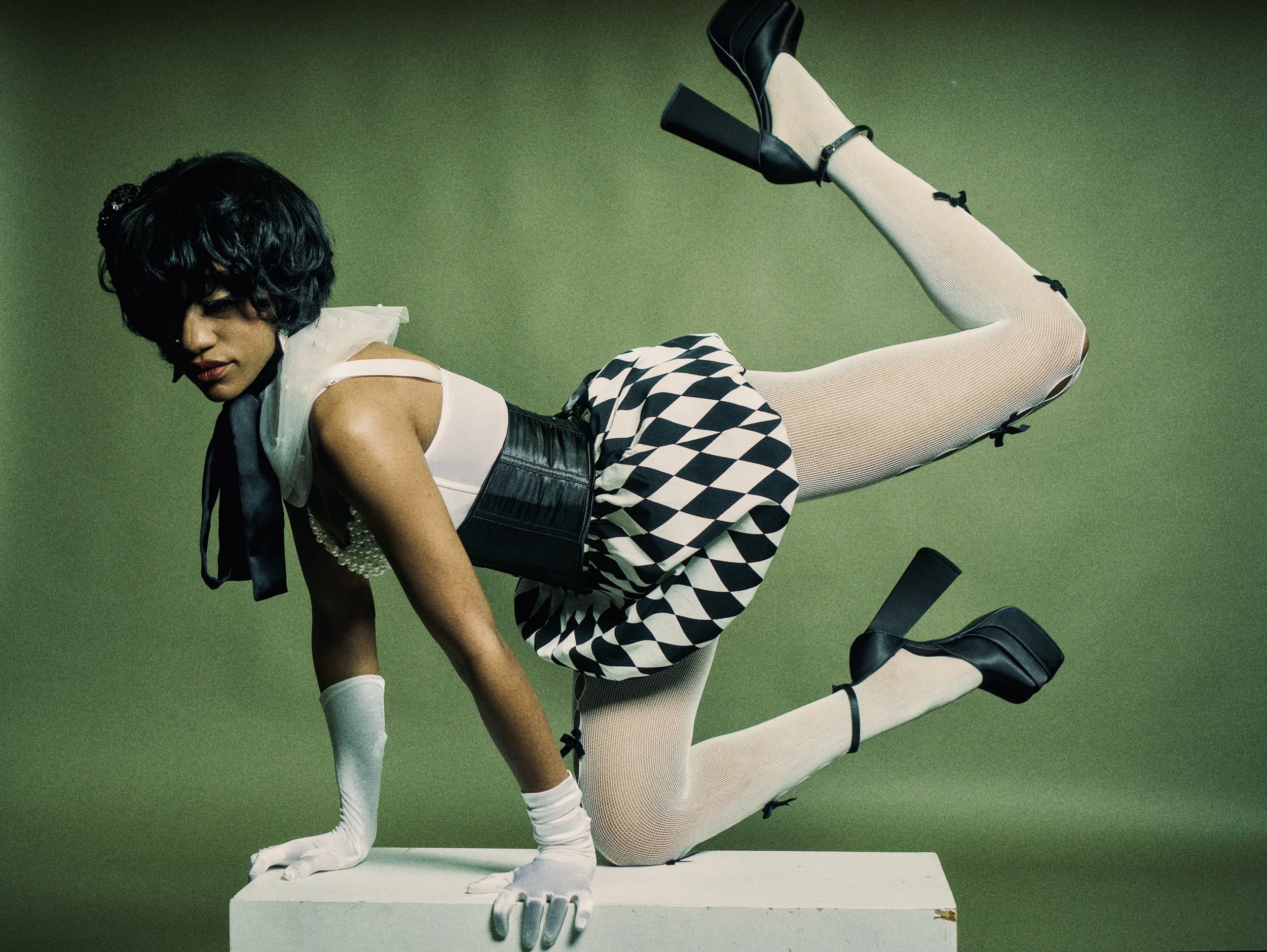 A woman with short black curly hair poses on a white pedestal against a green background. She is dressed in a white top, black corset, checkered skirt, white fishnet stockings, and black high-heeled shoes. She wears white gloves, a pearl necklace, an