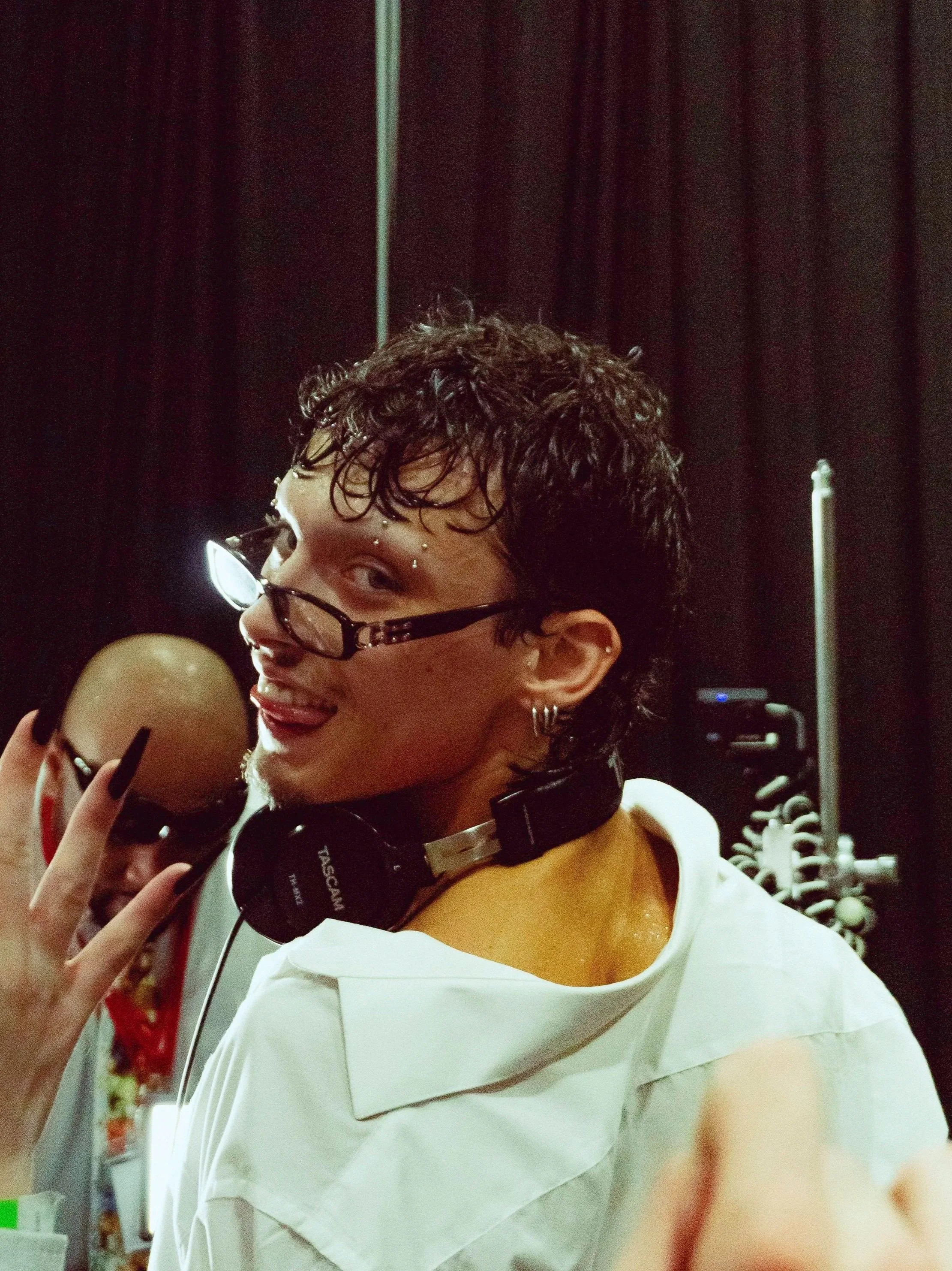 A person with short, curly hair, glasses, and multiple facial piercings, wearing headphones around their neck and a white shirt, making a playful expression while holding a mirror. This image of dj Connor Andre was taken by Erin De Shay.