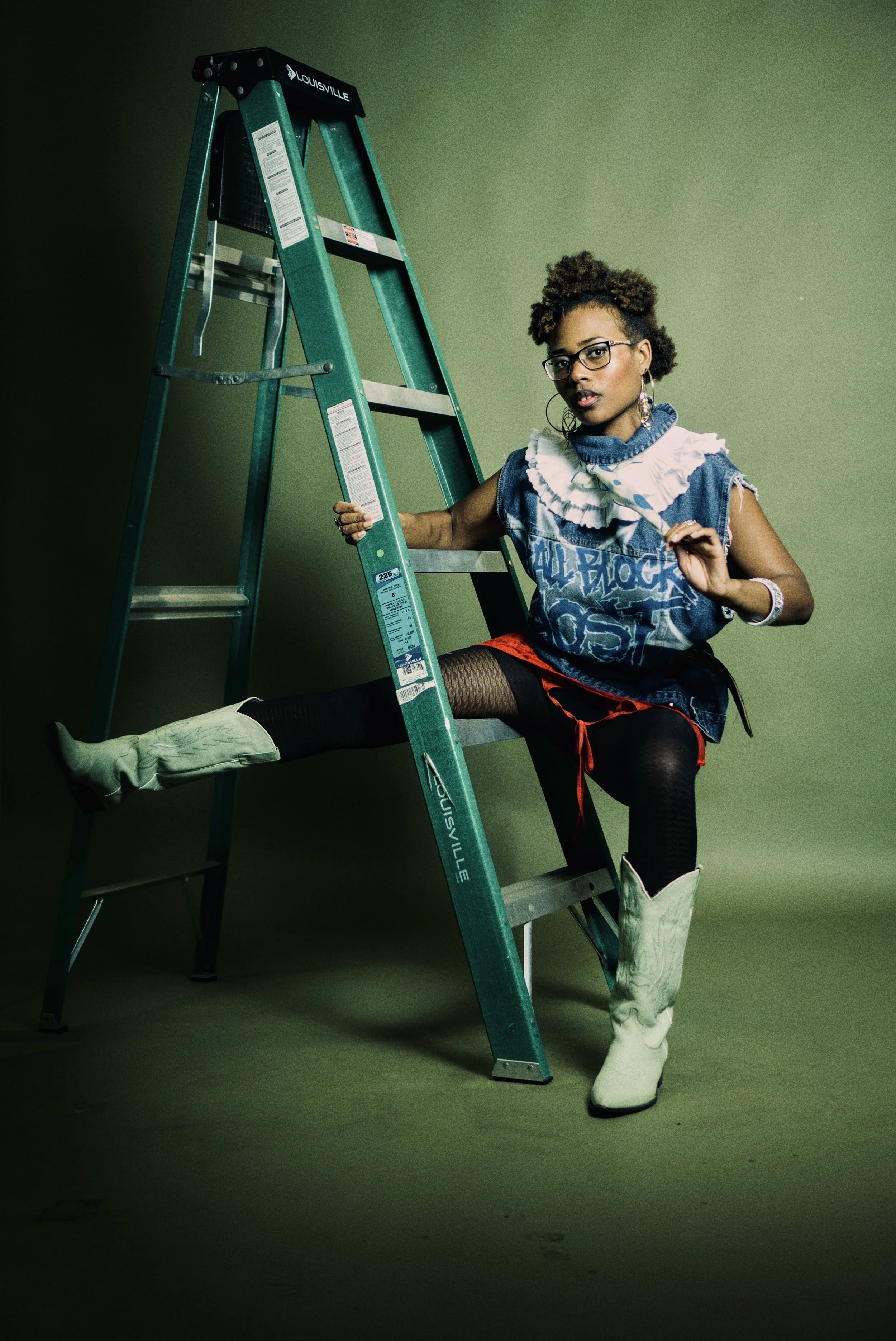 A woman with curly hair and glasses posing next to a green ladder against a plain green background, wearing a distressed graphic T-shirt, red shorts, fishnet stockings, and white cowboy boots.  Photographed by Erin De Shay.