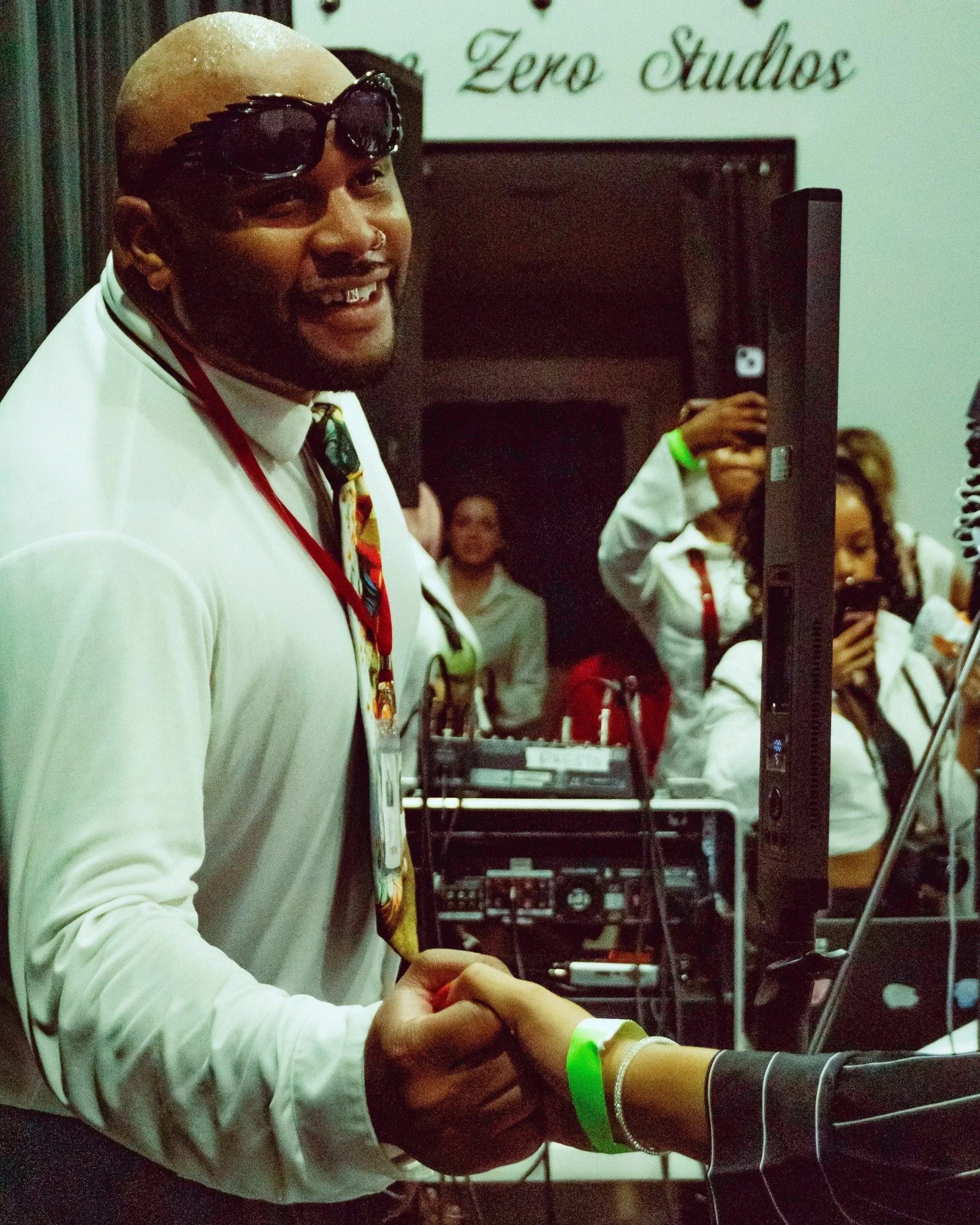 A man with a shaved head, wearing sunglasses on his forehead, a white shirt, and a colorful tie, shaking hands with someone. He has a grill in his mouth and a green wristband. In the background, there are people, some taking photos or videos, and a sign that says 'Zero Studios'.