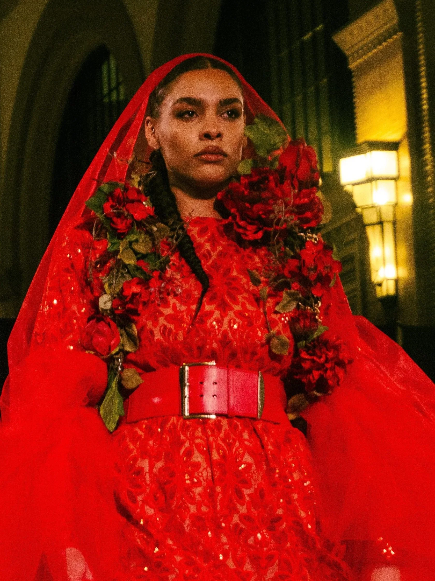 A woman dressed in an elaborate red outfit with floral details and a red veil, standing in a dimly lit indoor setting with warm lights during Richmond Fashion Week captured by fashion photographer Erin De Shay.