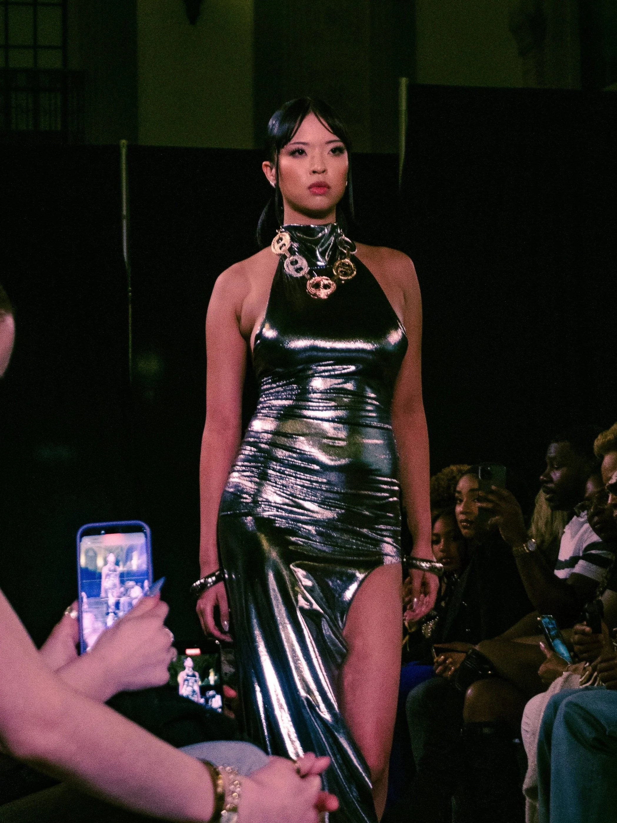 A model walking down a runway wearing a shiny black metallic dress with a high slit, accessorized with a chunky gold necklace and earrings, Richmond Fashion Week captured by fashion photographer Erin De Shay.