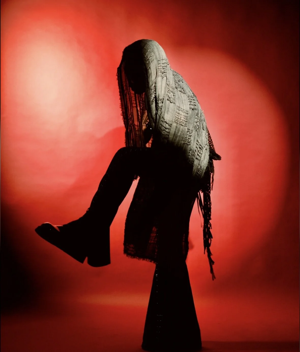 Silhouette of a man standing dramatically  with his head bowed, against a red background, wearing a long flare pants, lace top, and black boots.  Photographed by Erin De Shay.