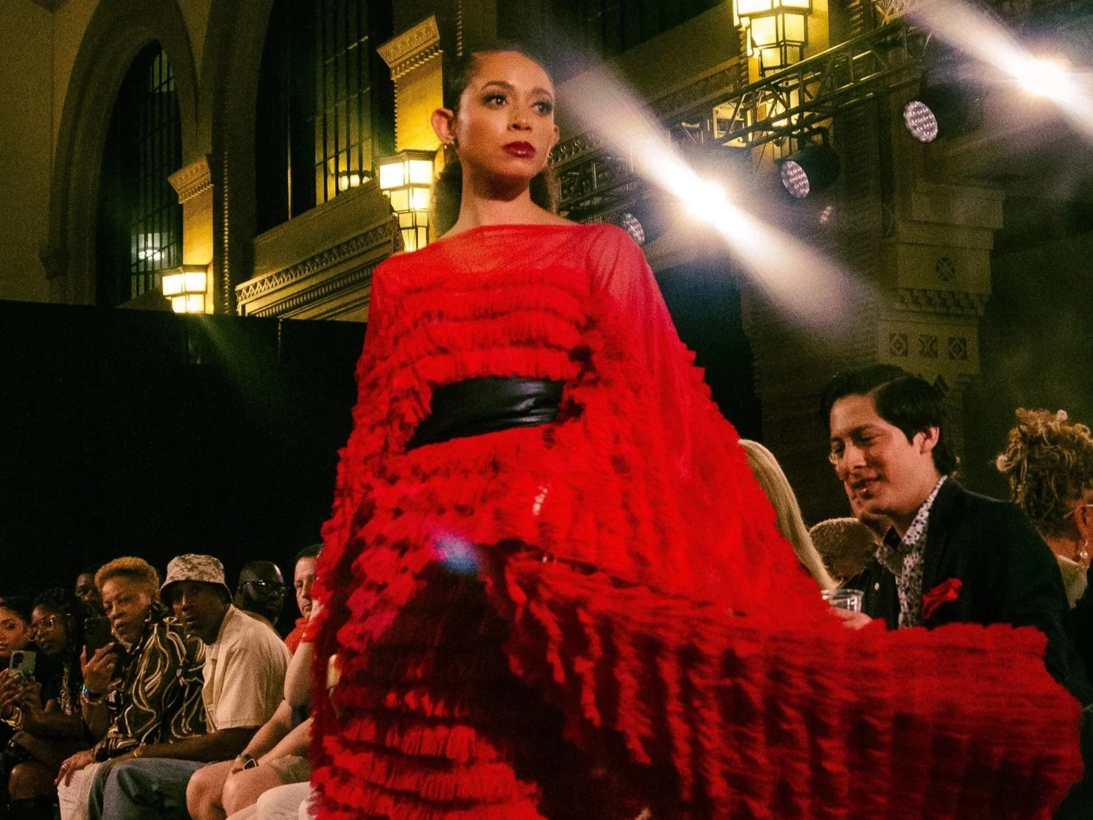 A woman in a vibrant red gown walking on a runway during a fashion show, with an audience seated on either side and a grand architectural background with large windows and ornate lighting  captured by fashion photographer Erin De Shay