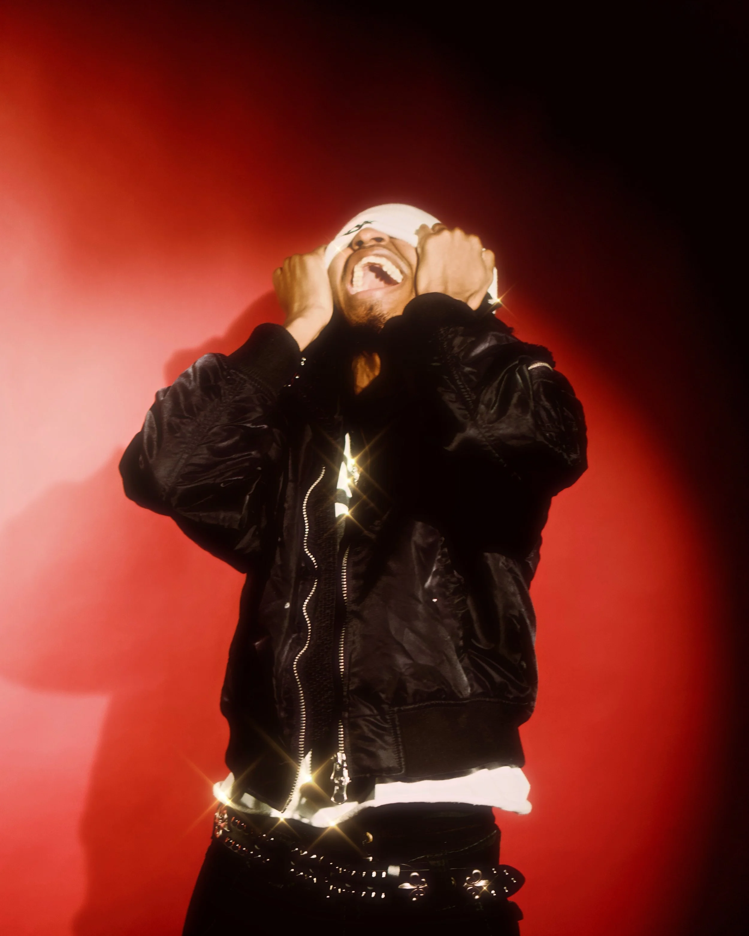 A man in a black jacket and white headwrap, laughing with eyes closed, against a red background, with sparkle effects.  Photographed by Erin De Shay.
