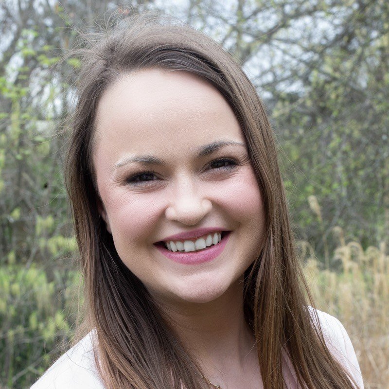 A young woman with long brown hair smiling outdoors in a natural setting with trees and plants in the background.