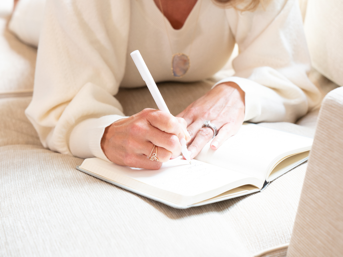 Person writing in a notebook with a white pen while seated on a beige couch.