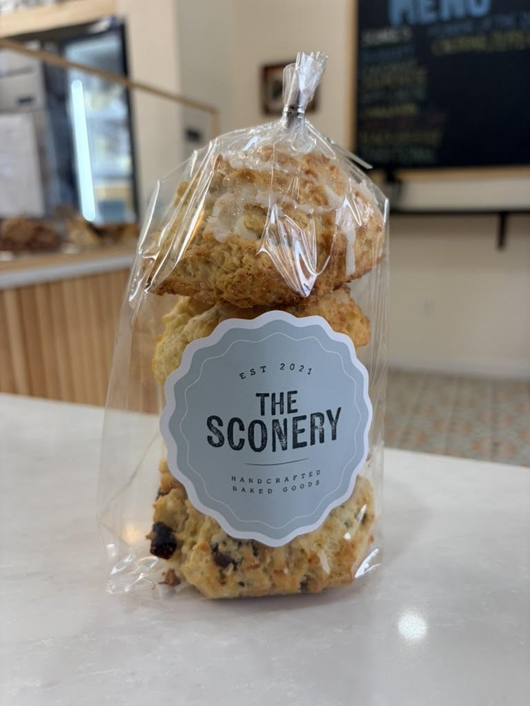 Clear plastic bag of assorted cookies with a label that reads 'The Sconery, Handcrafted Baked Goods, Est 2021' on a countertop inside a bakery or cafe.
