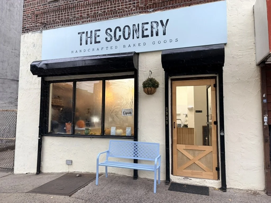 Exterior of a bakery called The Sconery with a light blue sign, black awning, and a wooden door, with a blue bench in front and a window displaying baked goods