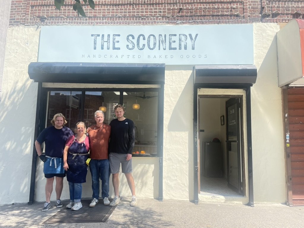 Four people standing outside of a bakery named 'The Sconery' with a sign that says 'Handcrafted Baked Goods'.