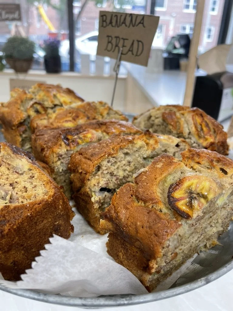 Banana bread slices with visible banana pieces in a basket lined with paper, with a handwritten sign labeled "Banana Bread" in the background.