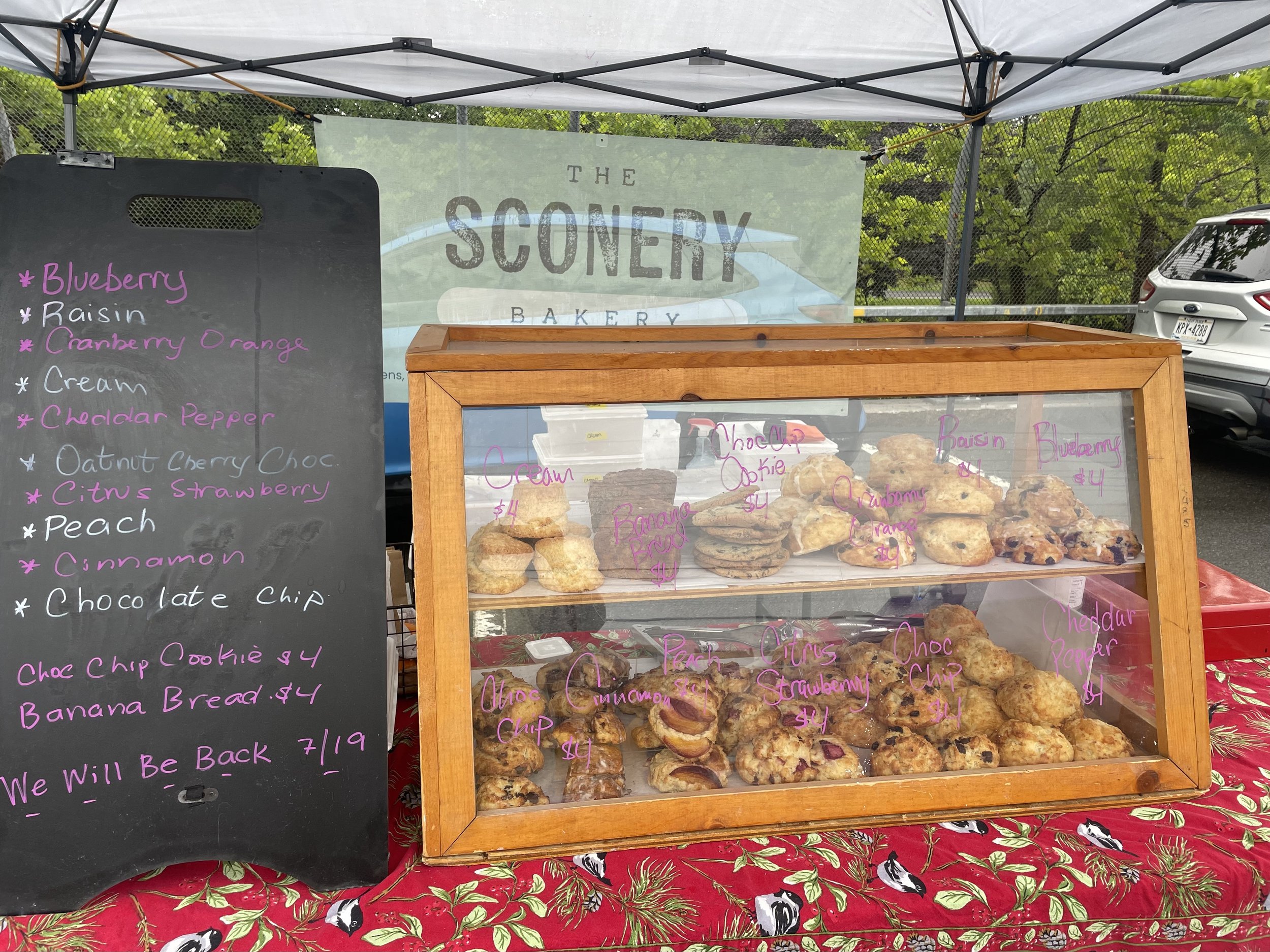 Display of assorted baked goods, including cookies and banana bread, at The Sonoma Bakery stand with a blackboard menu listing flavors, and a glass case showing individual cookies. The stand is outdoors with a tent, trees, and a parked car in the background.