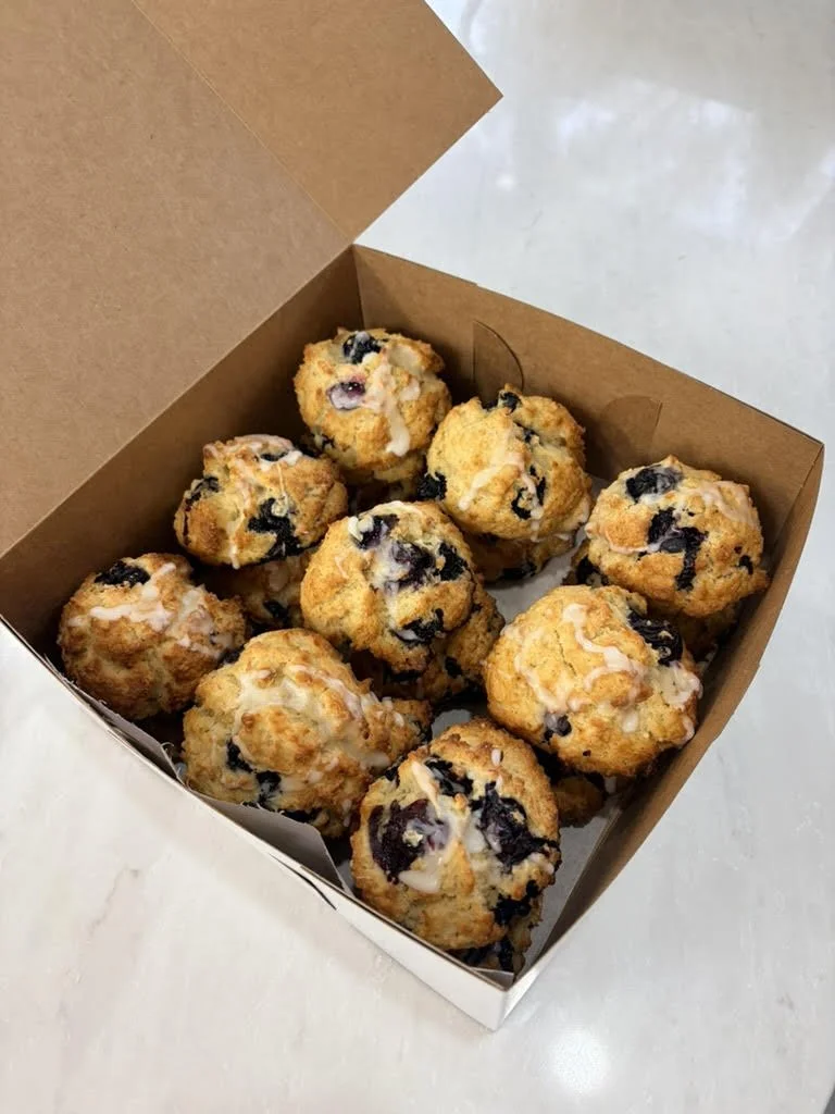 A cardboard box filled with blueberry muffins, some topped with white glaze or icing, sitting on a white surface.