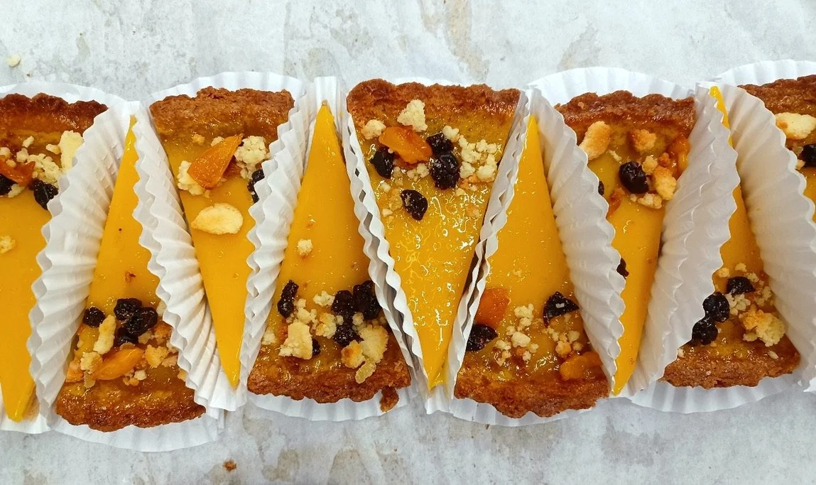Close-up of several slices of dessert with yellow custard, graham cracker crust, and toppings of fruit, crumbs, and chocolate chips, served in white paper cups.