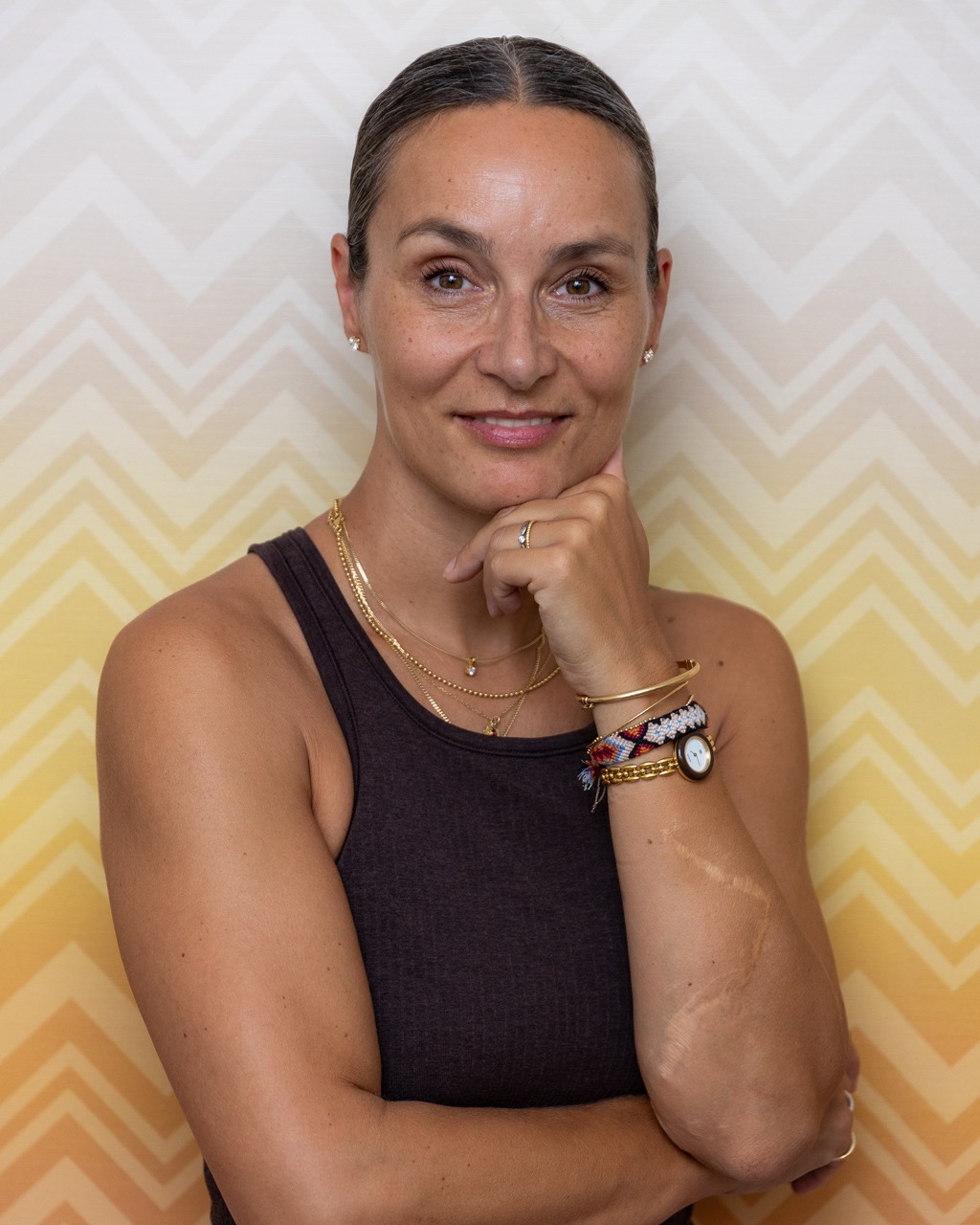 A woman with her hair pulled back, wearing a sleeveless black top, gold jewelry, and colorful bracelets, standing in front of a beige and yellow chevron patterned wall. She is resting her chin on her hand and smiling at the camera.