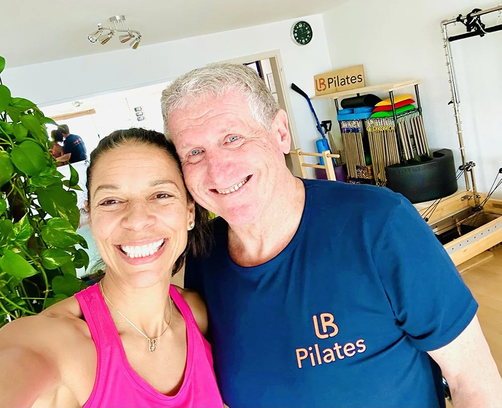 A smiling woman in a pink workout tank top and a man in a blue t-shirt with 'LB Pilates' logo take a selfie in a fitness studio. The background shows exercise equipment and a clock on the wall.