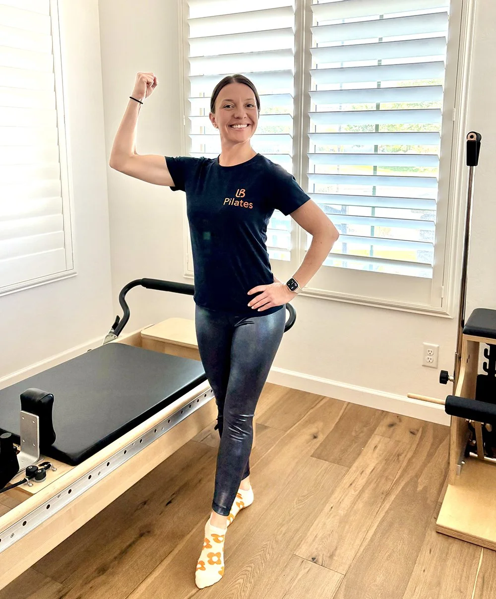 A woman in workout clothes, wearing a black Pilates shirt, shiny leggings, and white socks with orange flowers, is posing and flexing her right arm in a bright room with wooden floors and white plantation shutters.