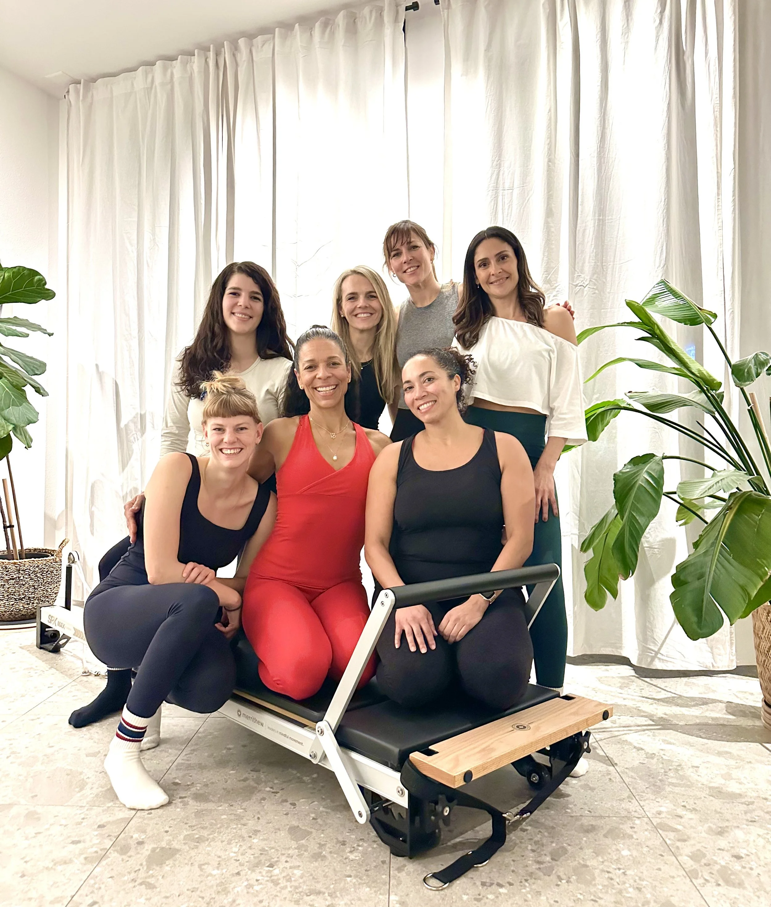 Group of eight women in a room with white curtains and large green plants, some standing and some sitting around a portable piece of exercise equipment.