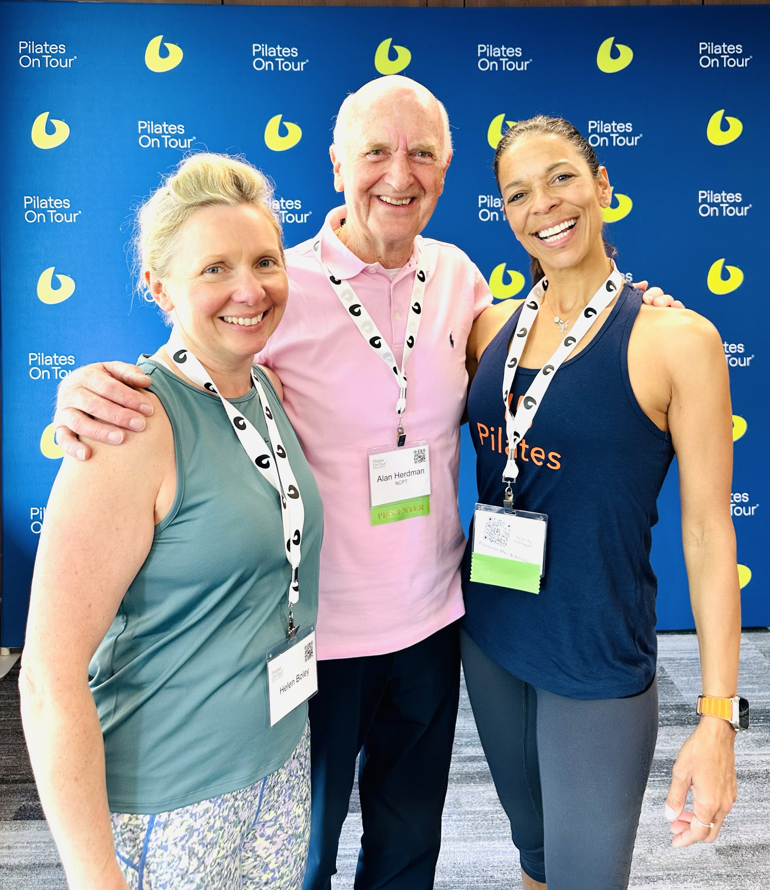 Three smiling people, two women and one man, standing together at a Pilates event, wearing conference badges, with a blue background featuring the Pilates On Tour logo.