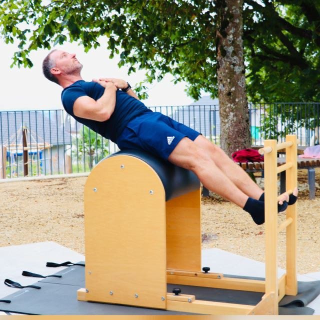 A man in a navy t-shirt and shorts performing a fitness exercise on a wood and metal aligned bench outdoors with green trees and a fence in the background.