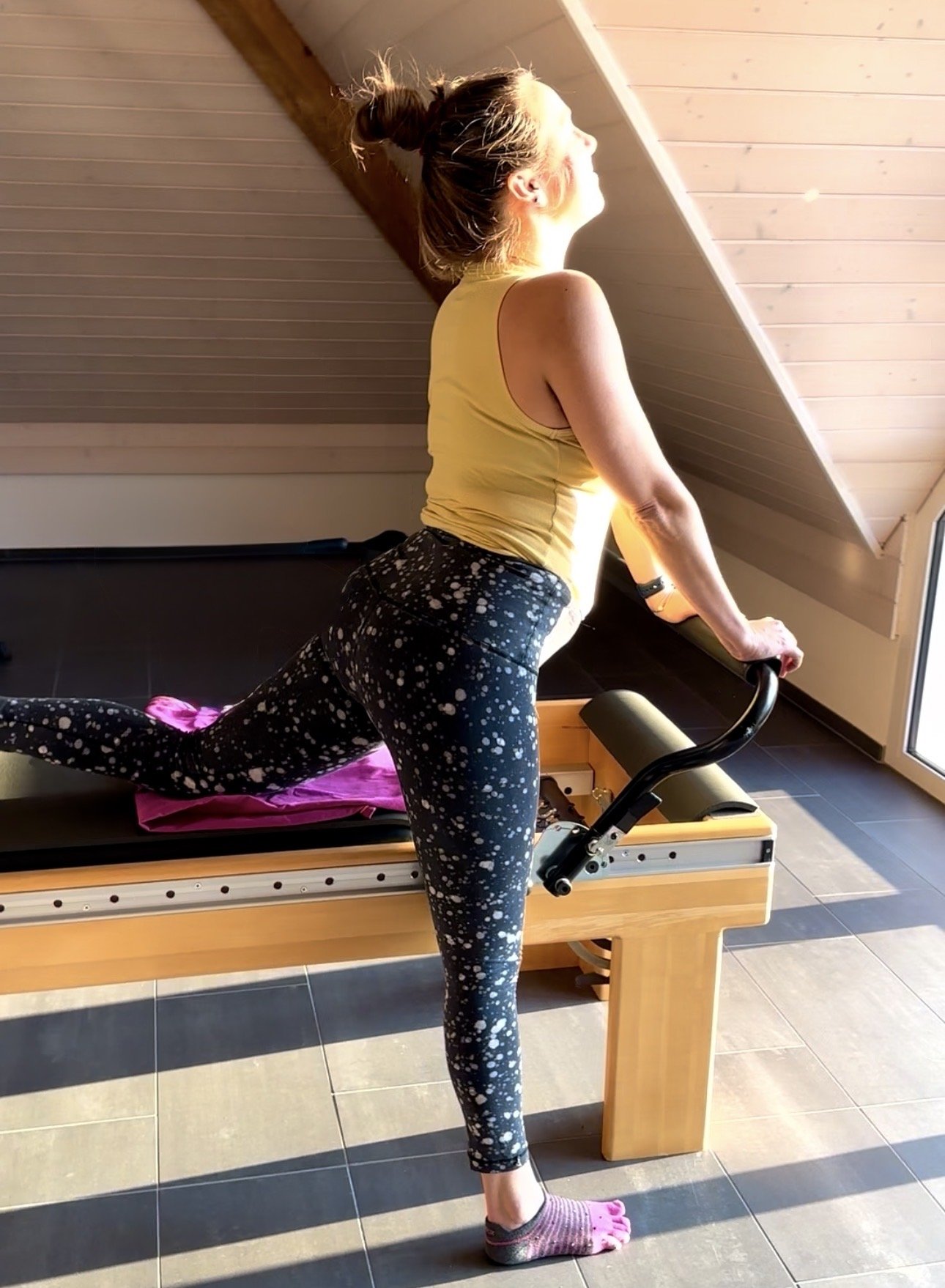 A woman with her hair in a bun, wearing a yellow tank top and black leggings with white splatter pattern, is performing a stretch with one foot elevated on a Pilates reformer machine in a room with wooden ceiling and floor tiles.
