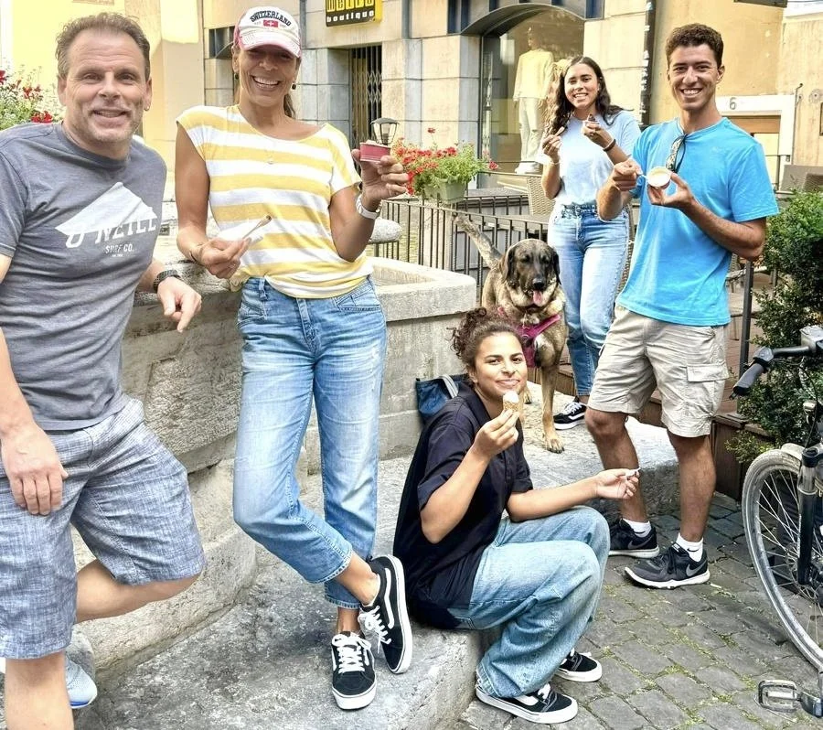 LaBriece family and a dog enjoying ice cream outdoors on a city street. They are smiling and holding ice cream cones, with some sitting on a stone ledge and others standing.