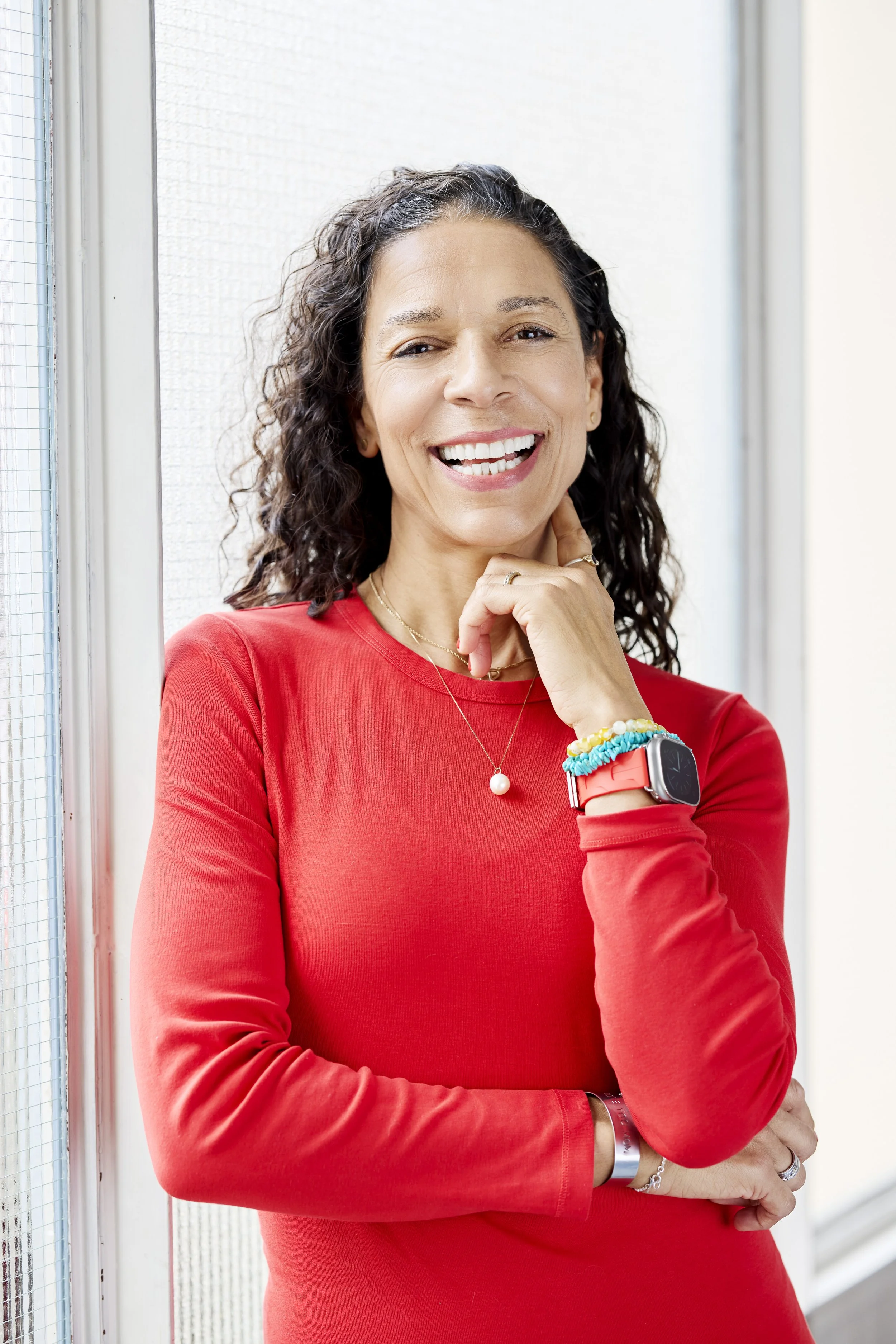 LaBriece smiling, wearing a red shirt, accessories, and a smartwatch, standing near a window.