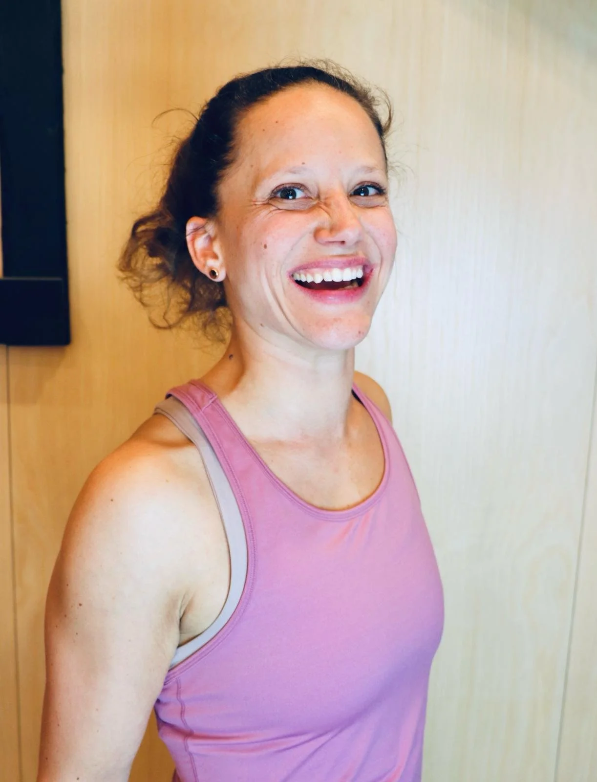 A woman in a pink tank top smiling and looking at the camera, with a light-colored wooden wall in the background.