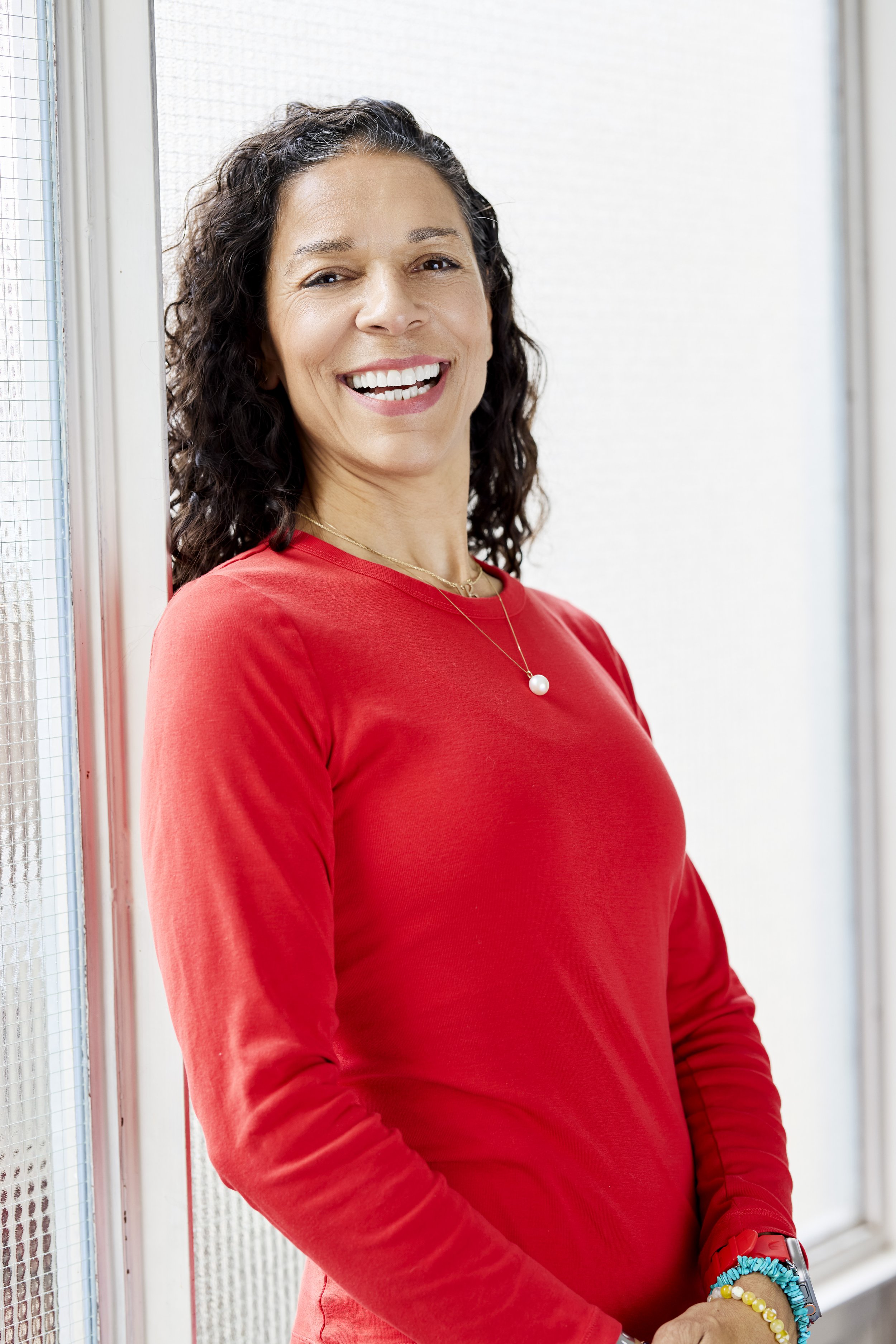 LaBriece smiling, wearing a red long-sleeve shirt and jewelry, standing by a window.