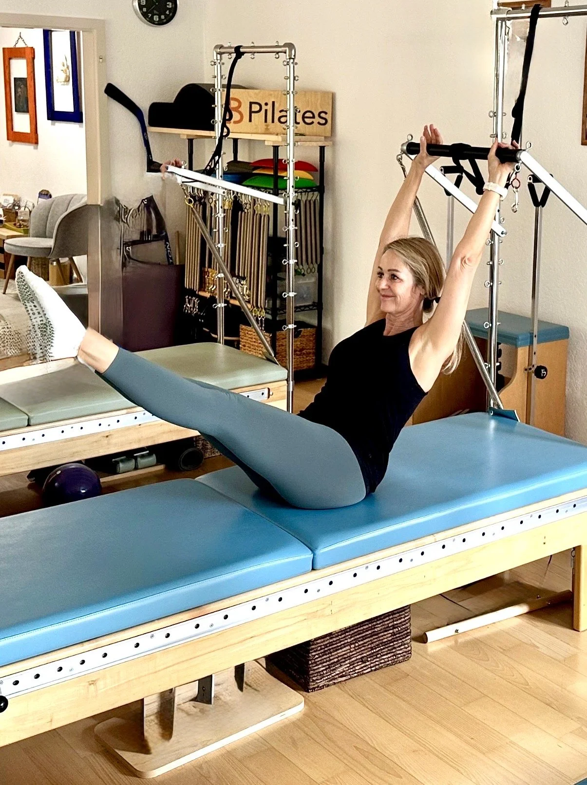 A woman doing a Pilates exercise on a reformer machine, lying on her back with her legs extended at an angle and holding onto the overhead bar with her arms raised.
