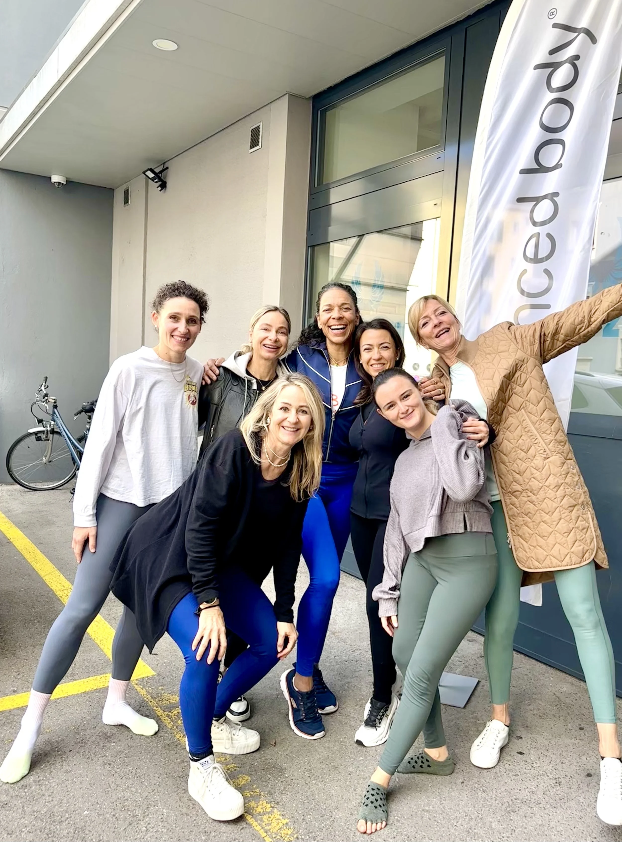 A group of seven women posing for a photo outside a building with a large vertical banner that reads 'Balanced Body'. Some women are smiling, one has her arm outstretched, and they appear to be casual or fitness attire.