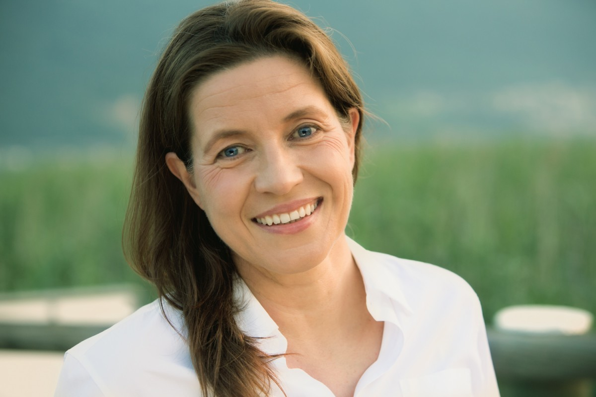 Close-up of a smiling woman with brown hair and blue eyes, wearing a white shirt, outdoors with a blurred green landscape background.