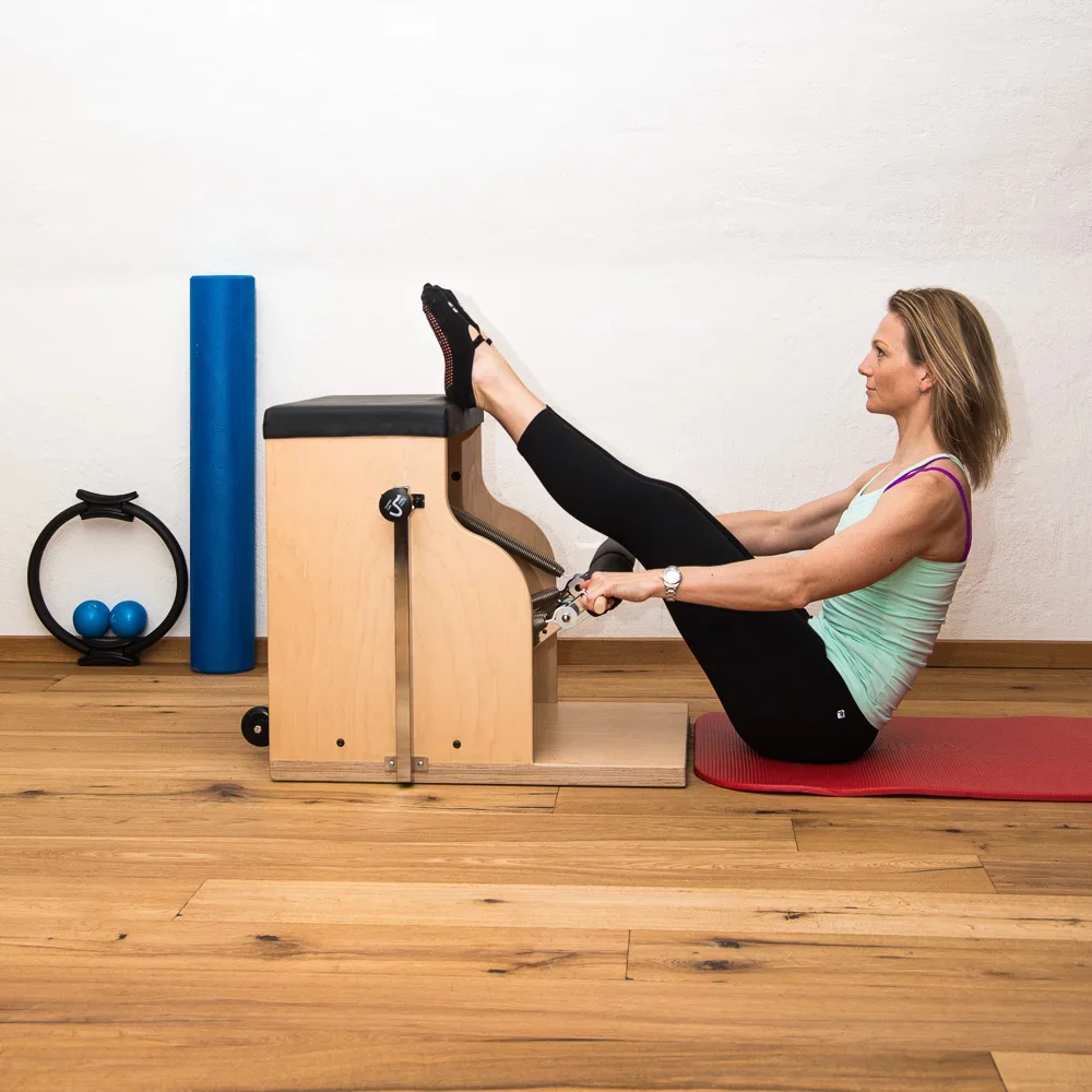 Woman exercising on a Pilates Reformer machine, wearing black leggings and a green tank top, on a red exercise mat in a room with wooden floors and fitness equipment in the background.