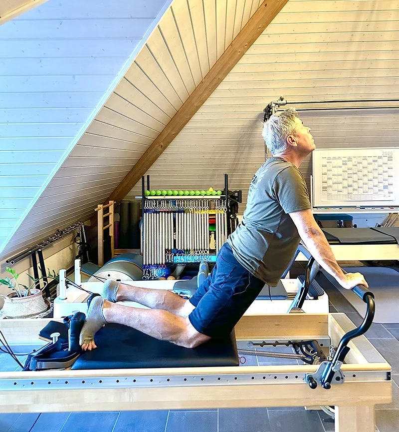A man exercising on a reformer Pilates machine in a home gym under a sloped ceiling, with fitness equipment and a calendar on the wall in the background.