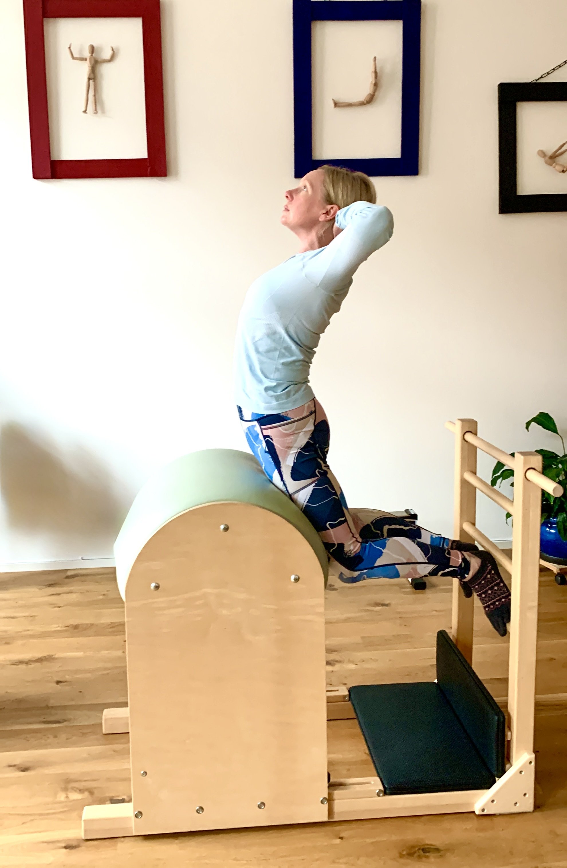 A woman stretching on a specialized piece of exercise equipment with a wooden frame and padded surface in a room with wooden flooring and artwork on the wall.