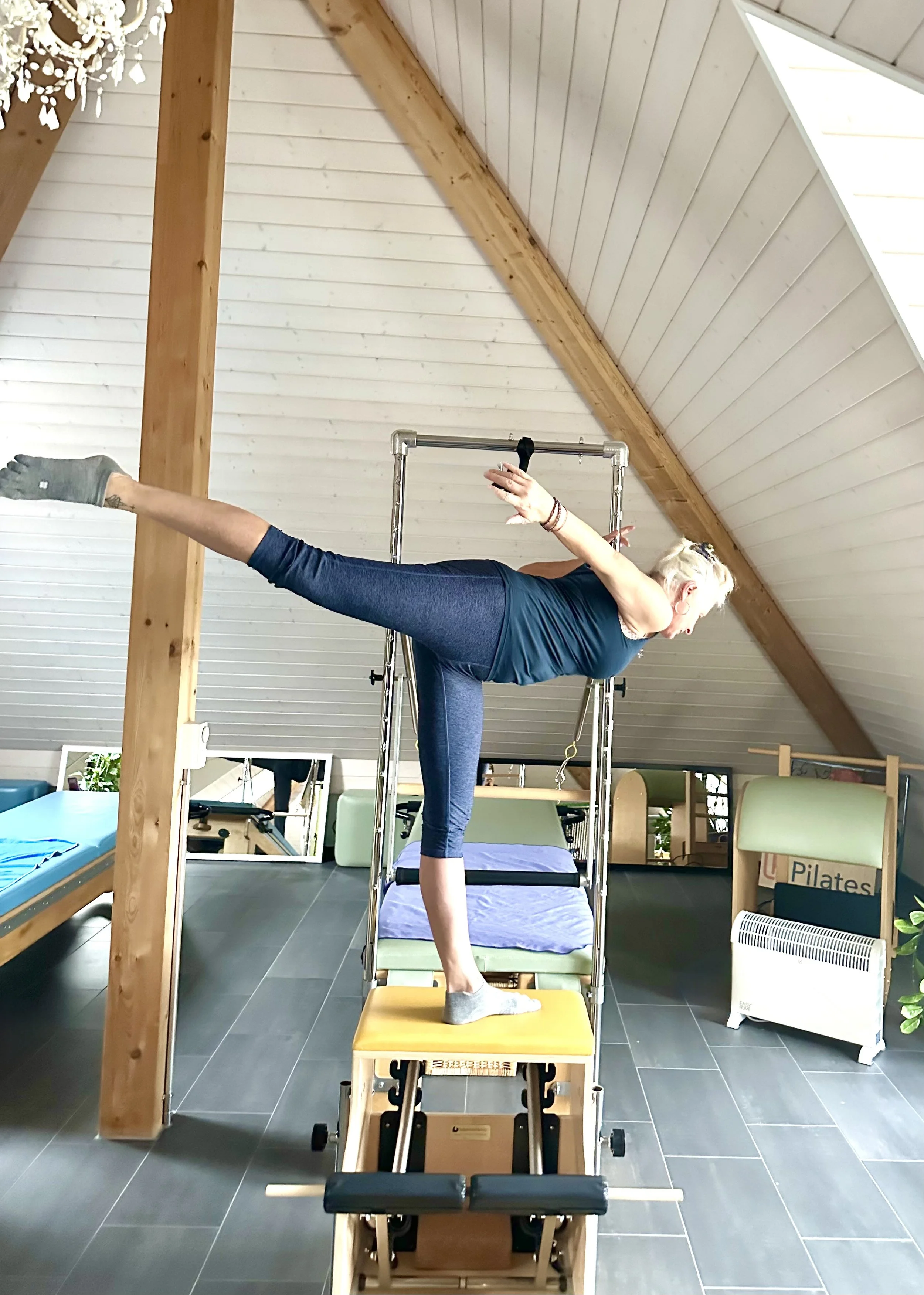 A woman performing a balancing exercise on Pilates equipment in a bright attic space with sloped ceiling, mirrors, and wooden beams.