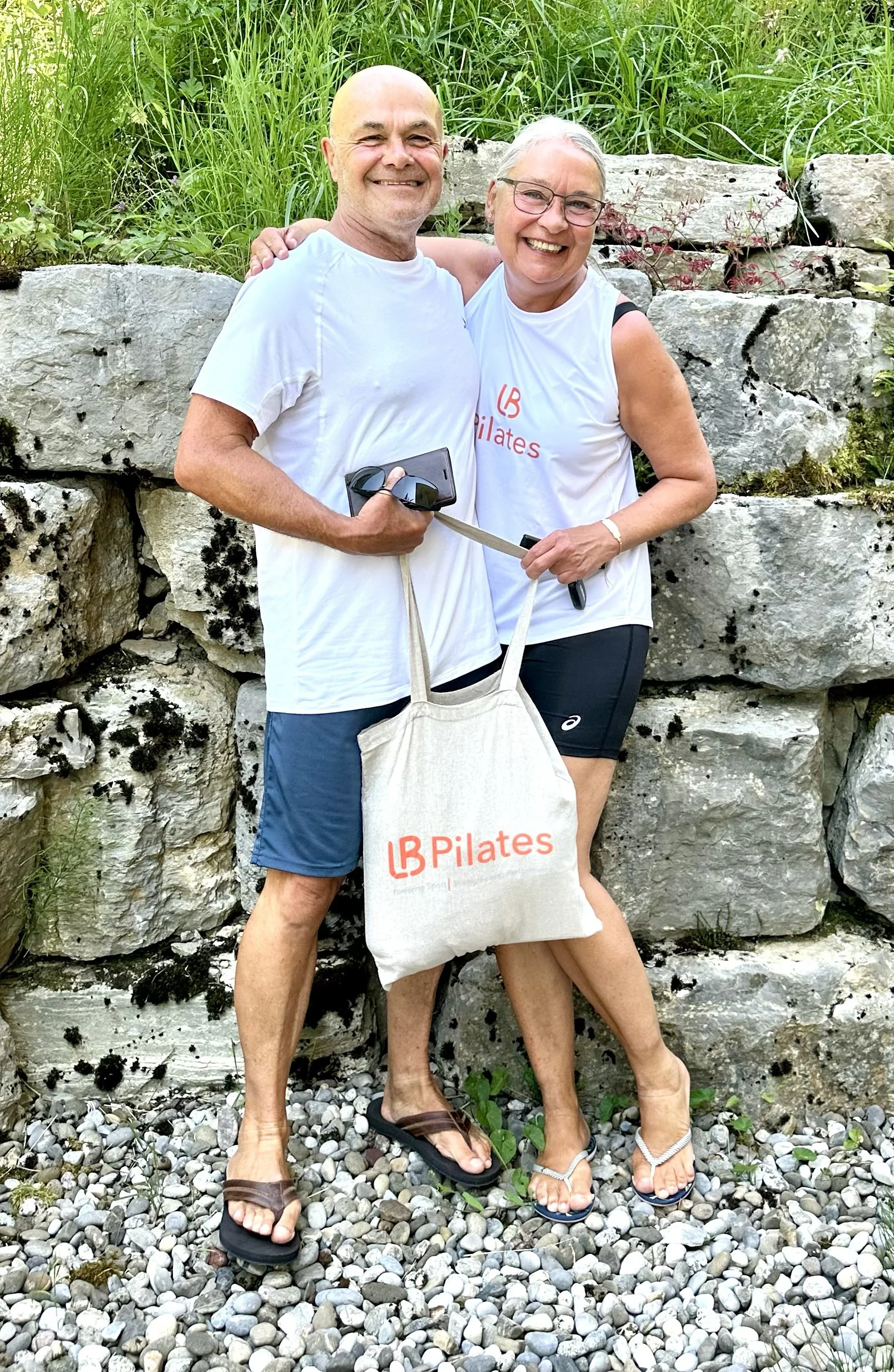 A smiling middle-aged man and woman stand close together outdoors in front of a stone wall and green grass, holding a tote bag with an "LB Pilates" logo, wearing casual summer clothing and sandals.