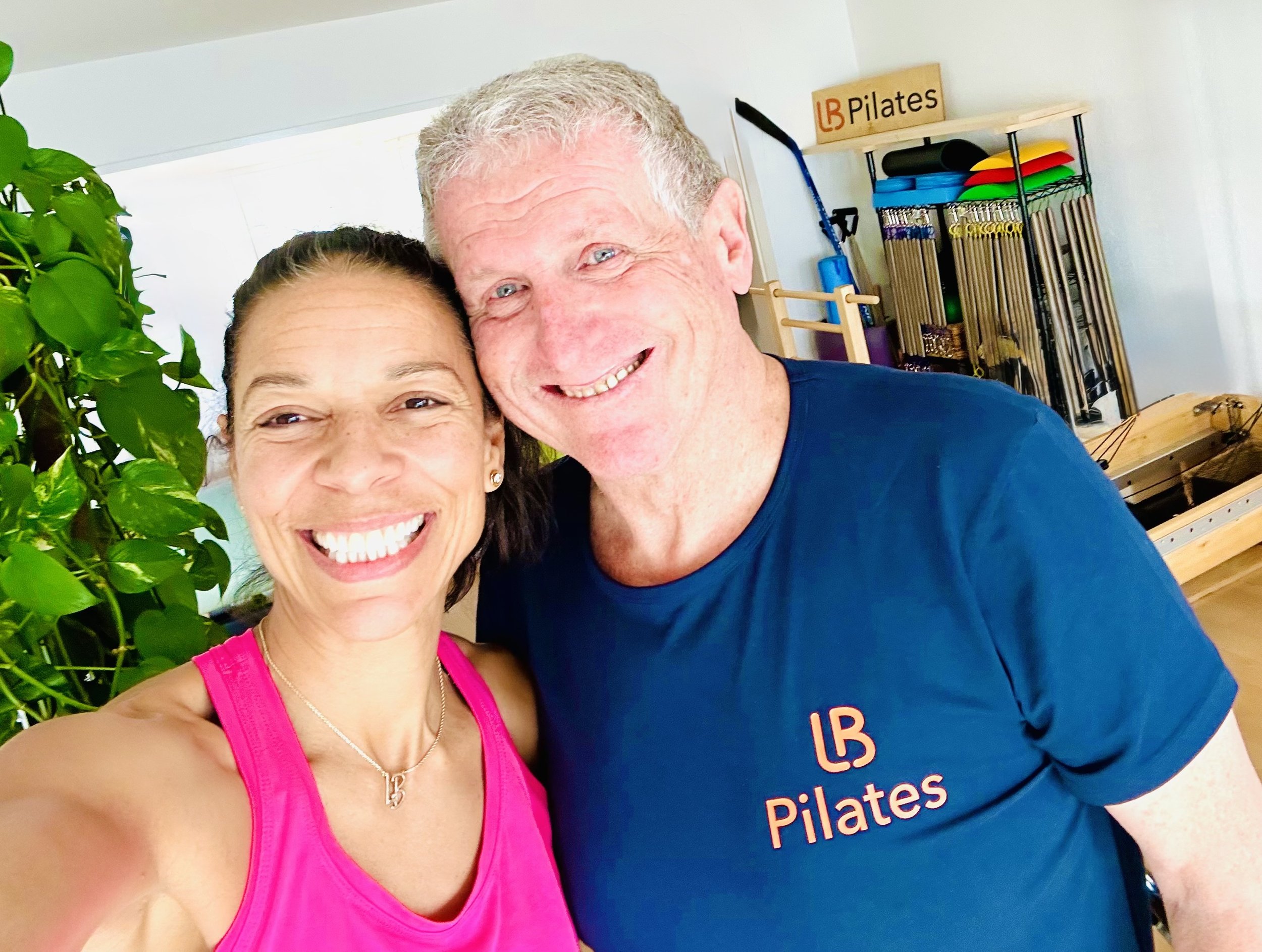 Smiling woman in pink fitness top and smiling man in Tom Pilates shirt taking a selfie together in a fitness studio with plants and exercise equipment in the background.