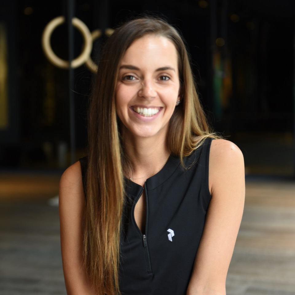A smiling young woman with long, light brown hair, wearing a black sleeveless athletic top, in a gym setting with fitness equipment in the background.