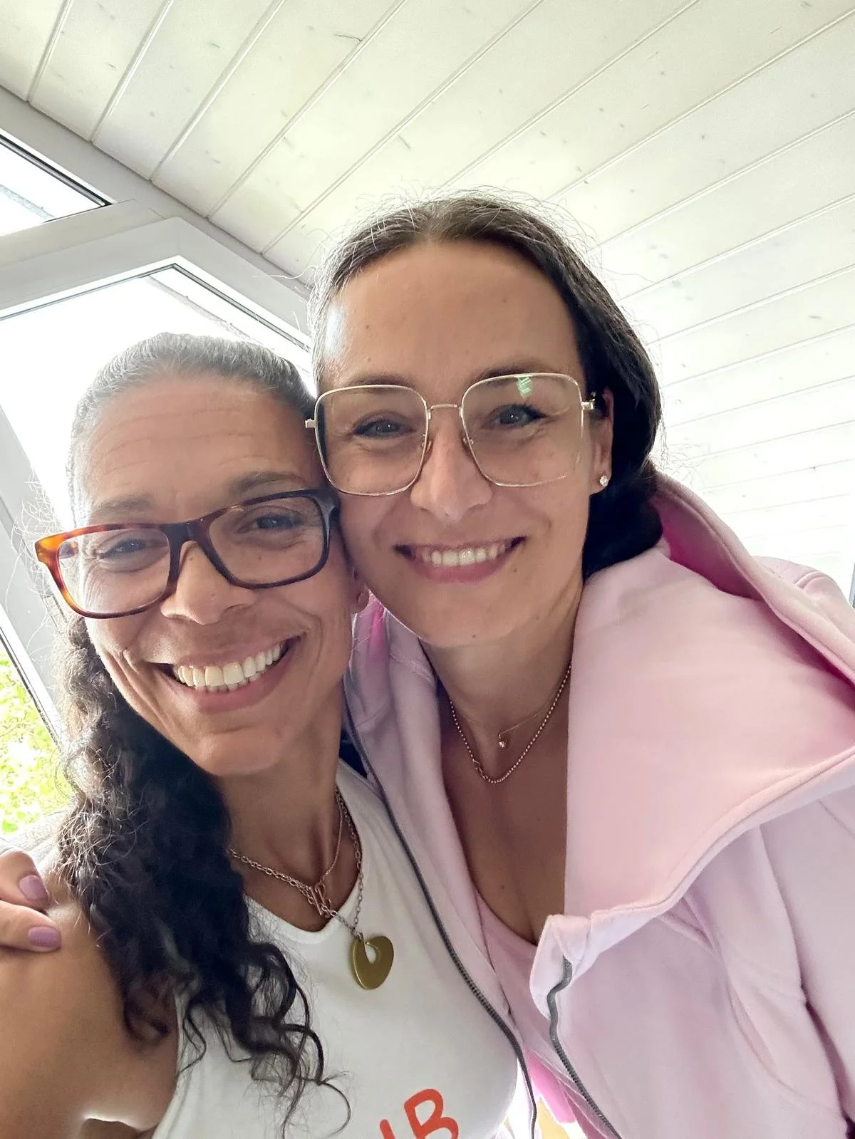 Two women smiling and taking a selfie indoors, with one woman wearing glasses and a white shirt, and the other woman wearing glasses and a pink blazer.